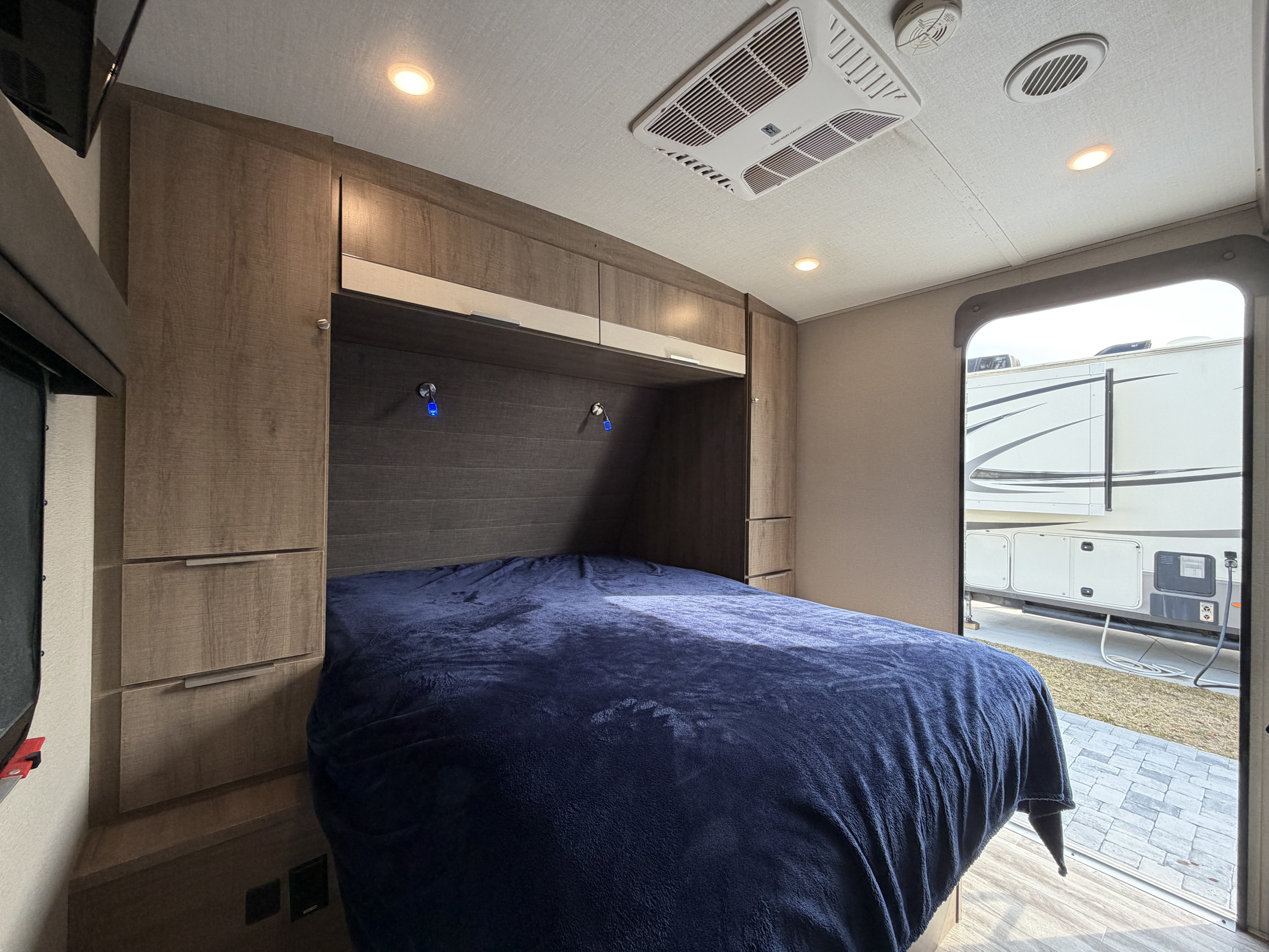RV rental interior: cozy bedroom with blue bedding, wood cabinets, overhead AC, and another RV visible outside.. Grand Design Imagine 2800BH #1 2021