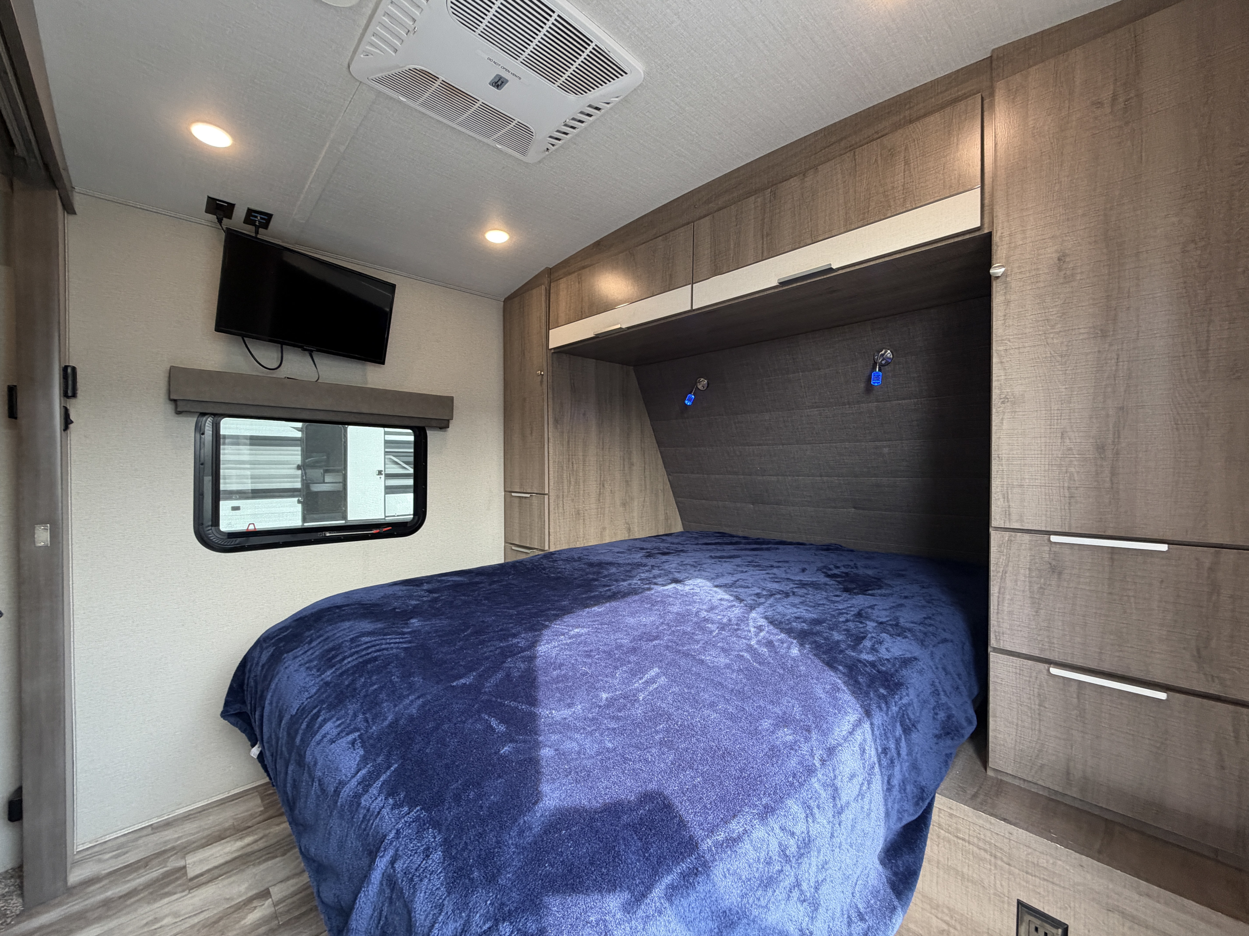 Cozy RV rental bedroom features a plush blue bed, wall-mounted TV, overhead cabinets, and a window for natural light.. Grand Design Imagine 2800BH #1 2021