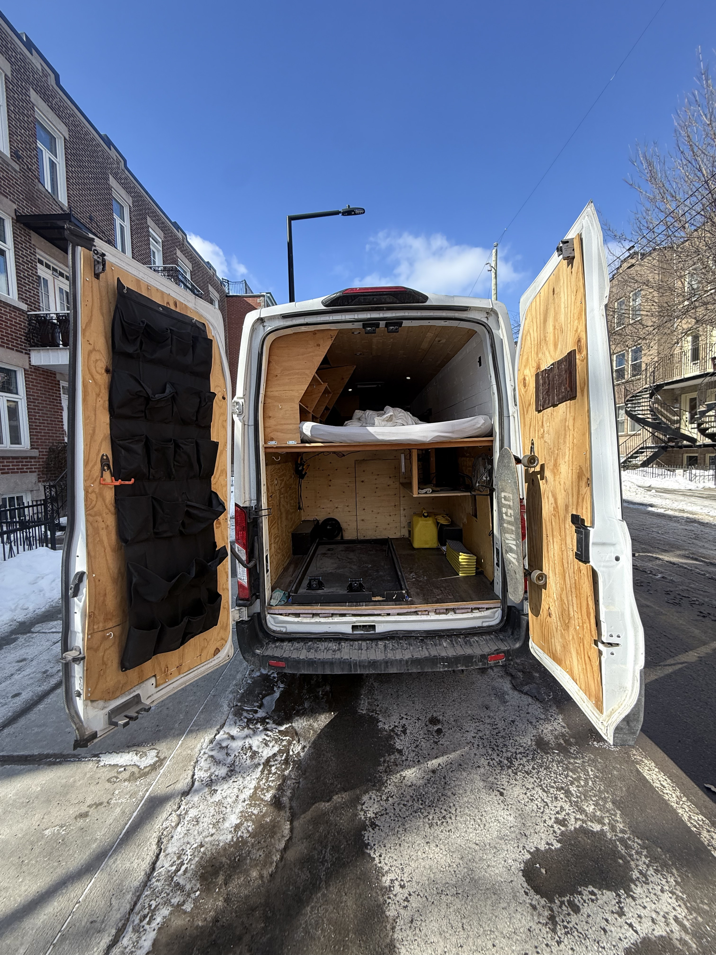 Inside an RV rental van: open rear doors reveal a custom wooden interior, bed, storage, and a skateboard, parked on a snowy street.. Ford Transit 2021