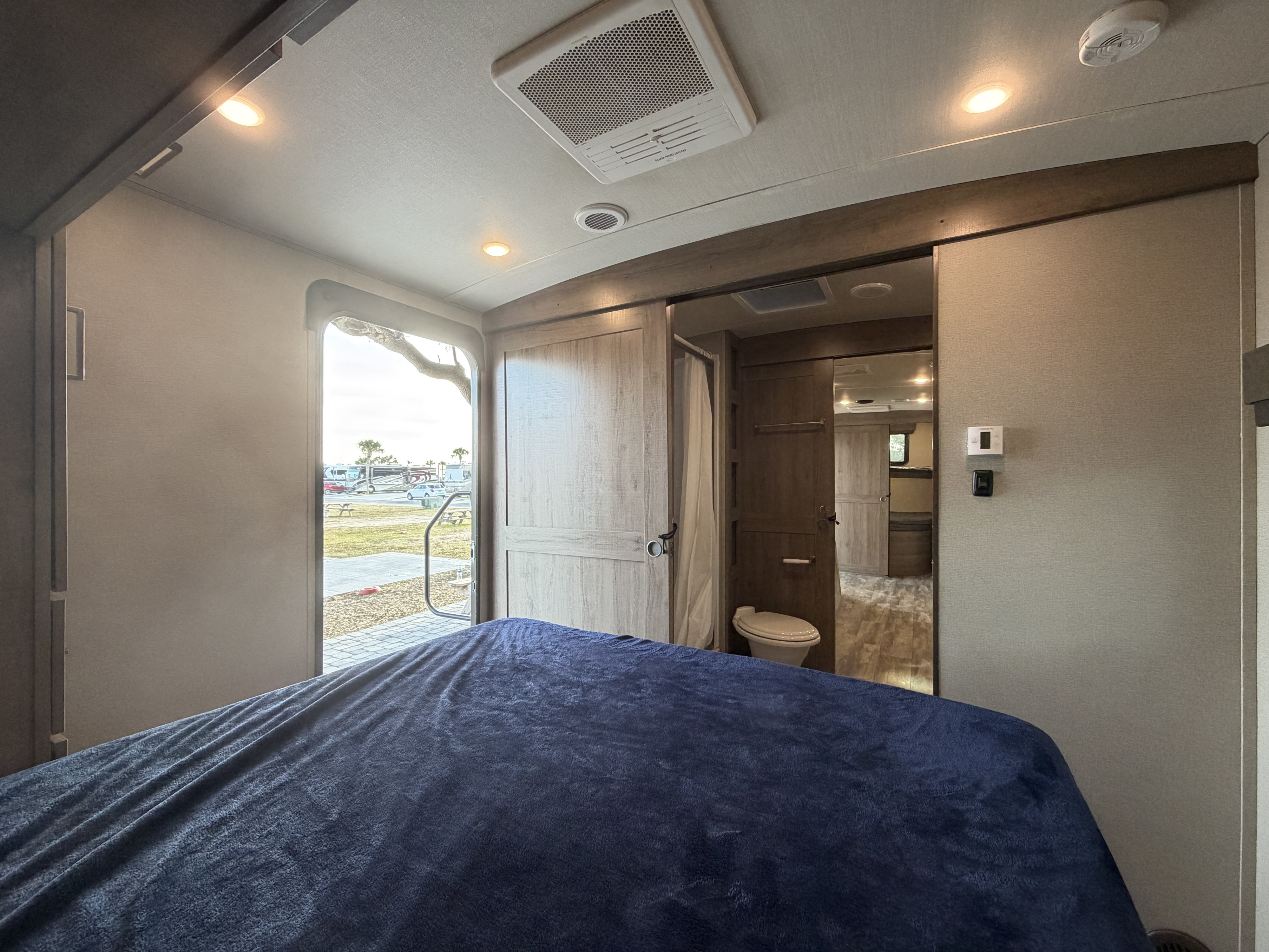 Inside an RV rental, this cozy bedroom features a blue bed, private bathroom, and an open door view of the campsite.. Grand Design Imagine 2022