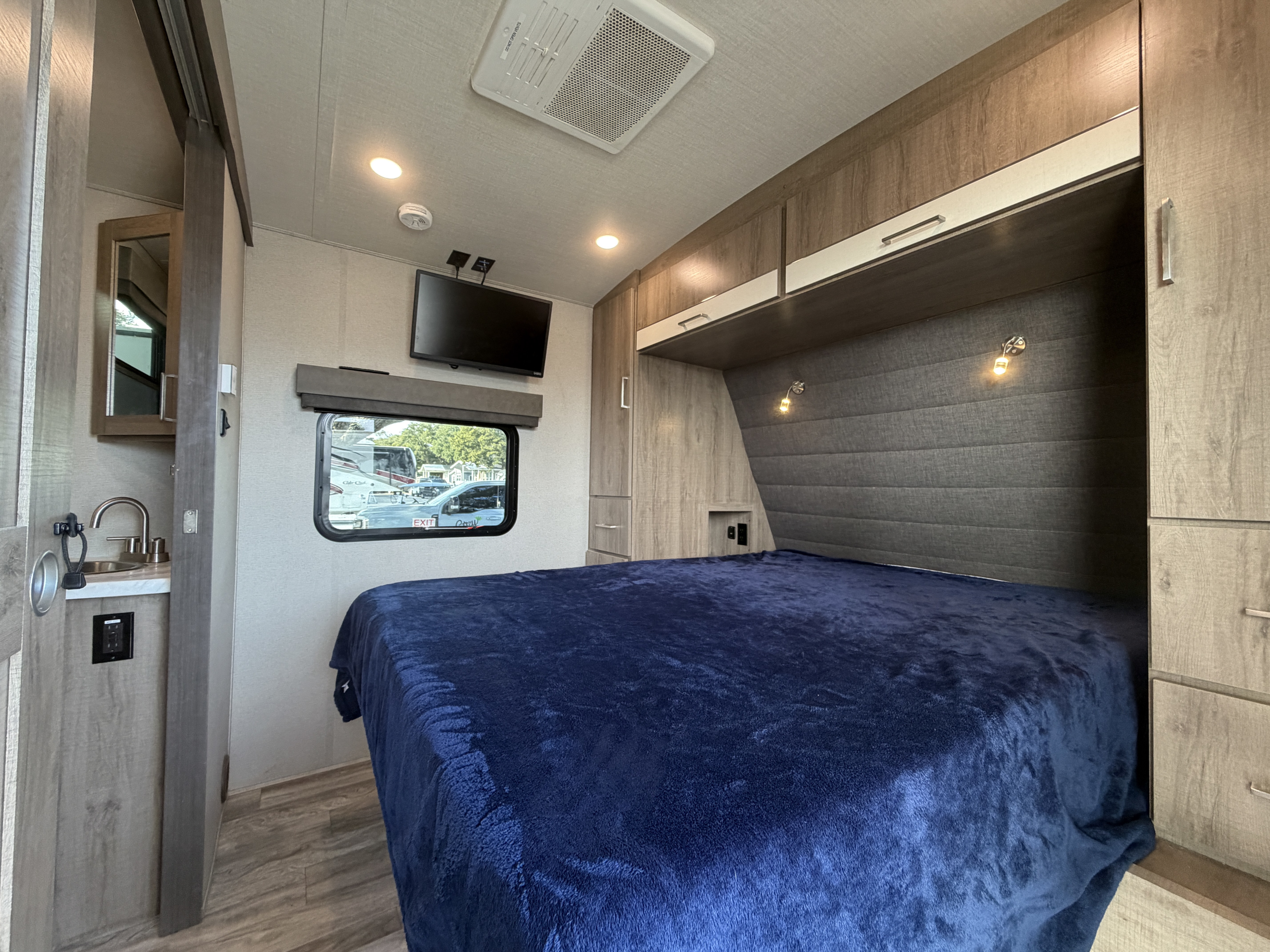 RV rental bedroom: a cozy space with a dark blue bed, TV, overhead cabinets, and a compact sink area.. Grand Design Imagine 2022