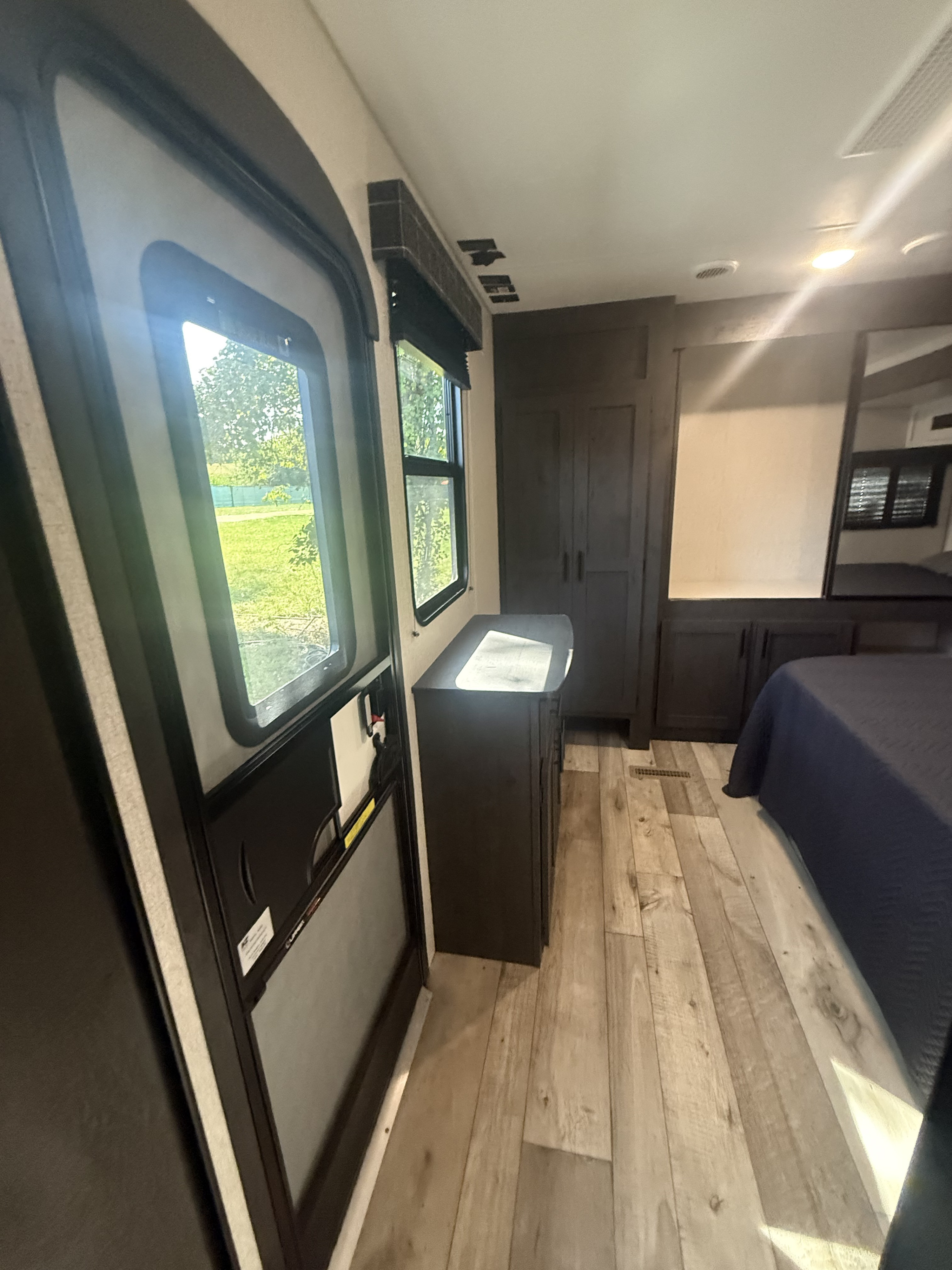 RV rental bedroom interior: dark wood cabinets, comfy bed, light wood floor, and sunlight through the entry door and window.. KZ RV Sportsmen 2022