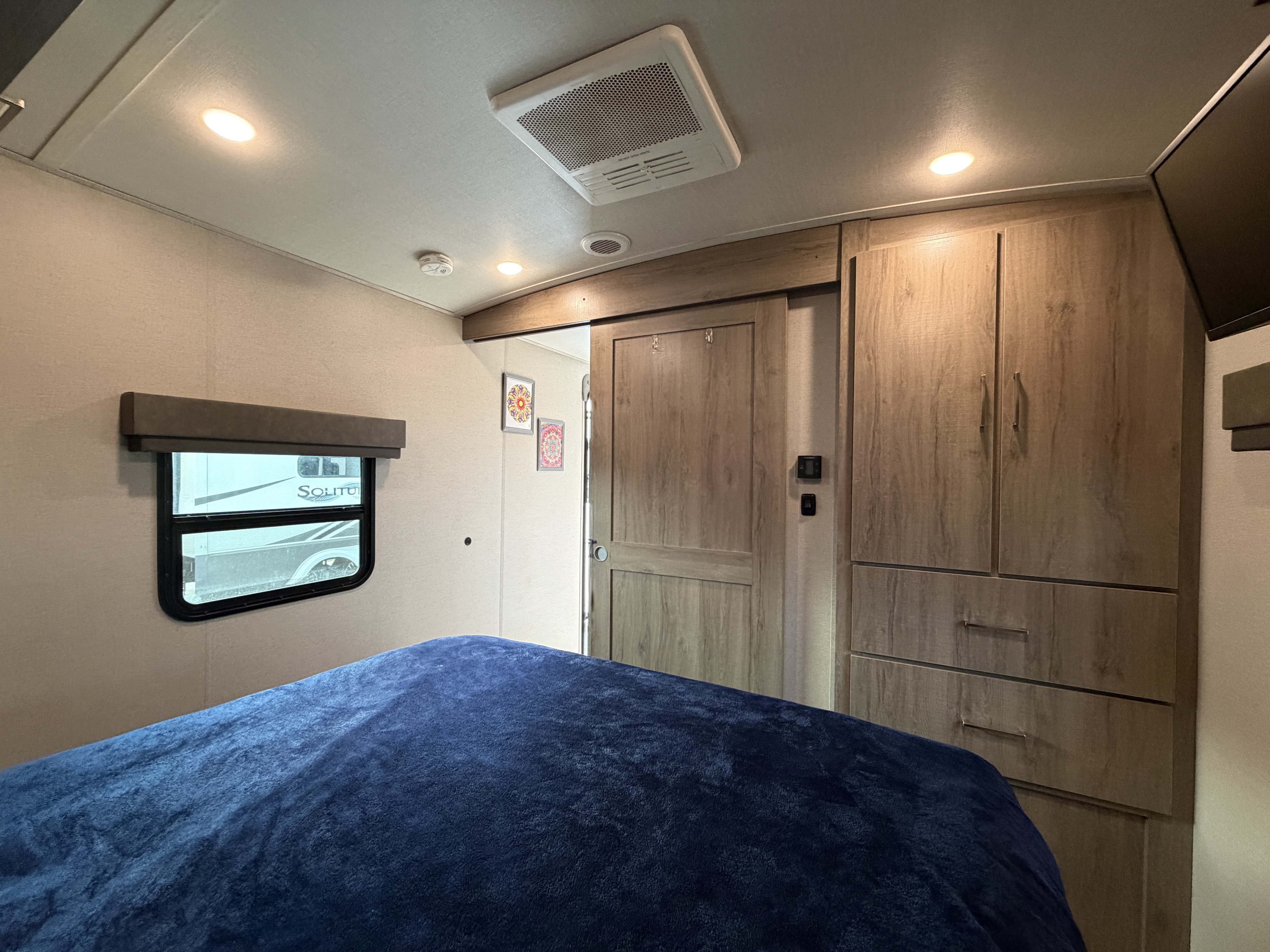 Cozy RV rental bedroom with a blue blanketed bed, wood cabinets, and a window, designed for comfortable mobile travel.. Grand Design Imagine 2023
