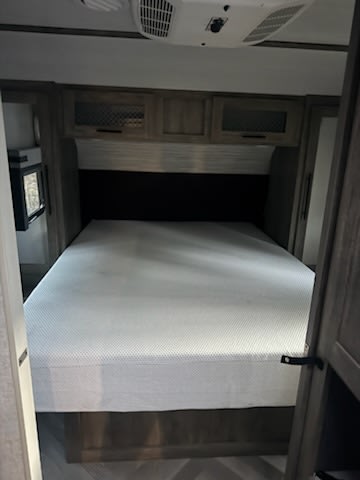 Inside this cozy rv rental, a comfortable bed with a white mattress cover, overhead cabinets, and a modern headboard.. Forest River Wildwood Lamitated Towables 2021
