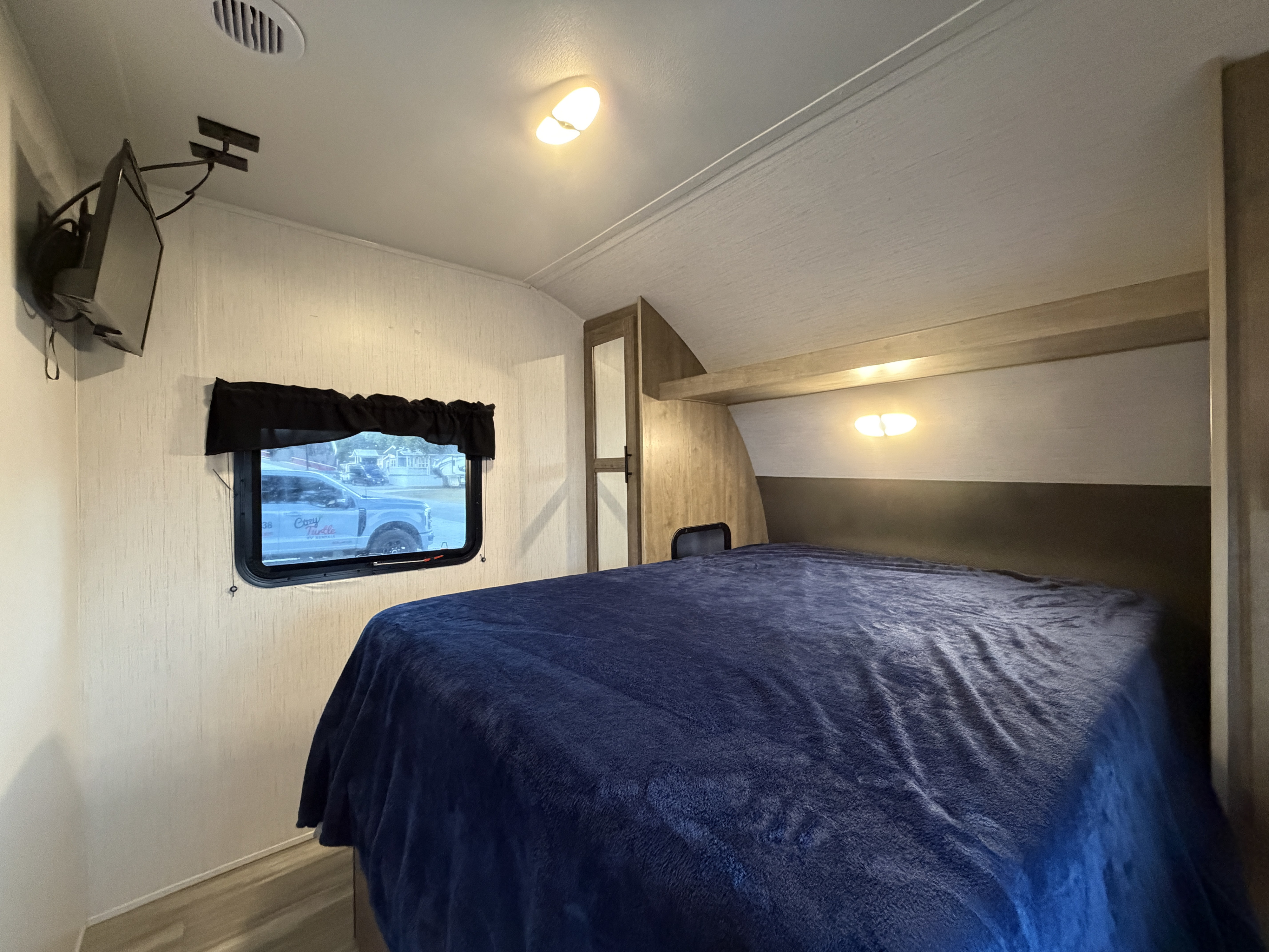 Cozy RV rental bedroom featuring a plush blue bed, wall-mounted TV, and window view of a "Cozy Turtle" truck.. Heartland Pioneer 2022
