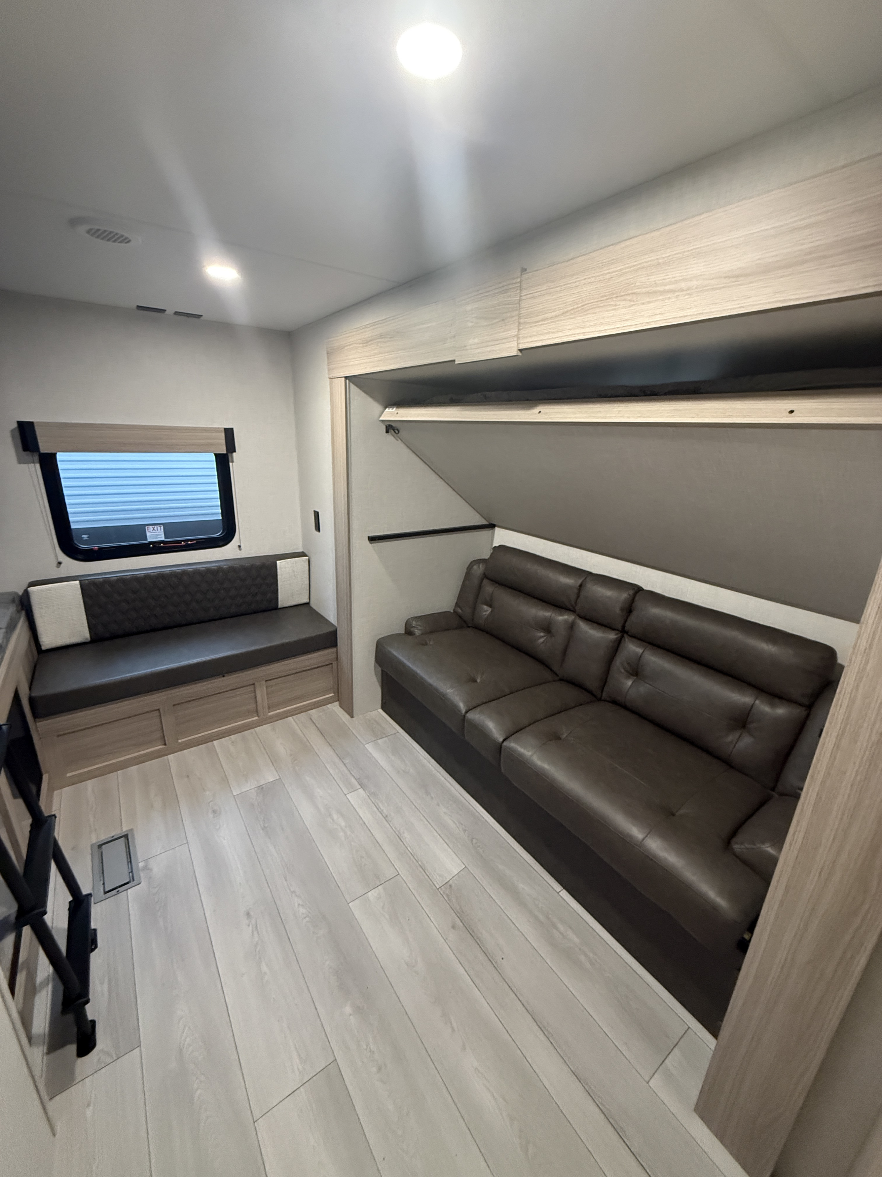 Clean RV rental interior featuring a dark brown sofa, patterned bench, light wood flooring, and an elevated bunk bed.. Kz Sportsmen 332bhkse 2026