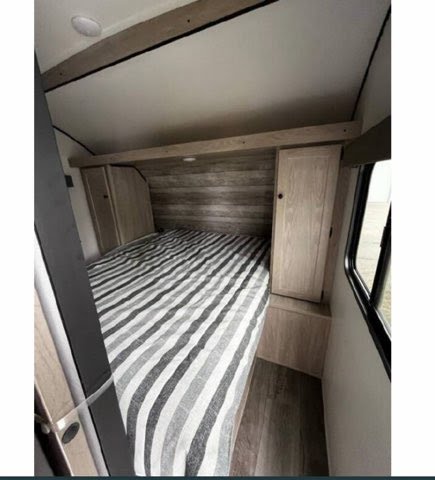 Inside an RV rental, a compact bedroom features a bed with striped bedding, built-in storage, wood accents, and a window.. Riverside RV Intrepid 2023