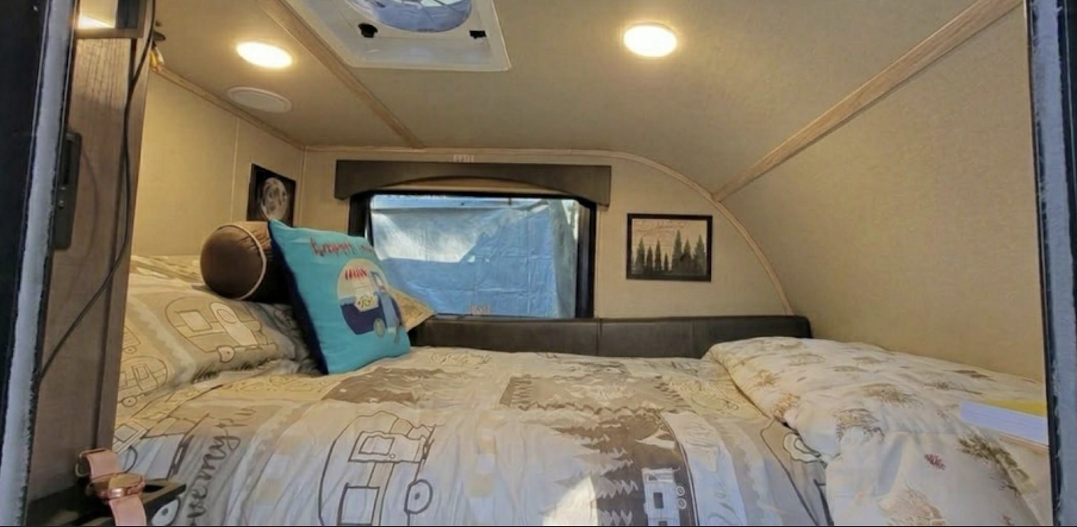 Cozy RV rental interior: bedroom with patterned bedding, blue 'Happy Camper' pillow, window, and ceiling fan.. Sunset Park RV SunRay 2024