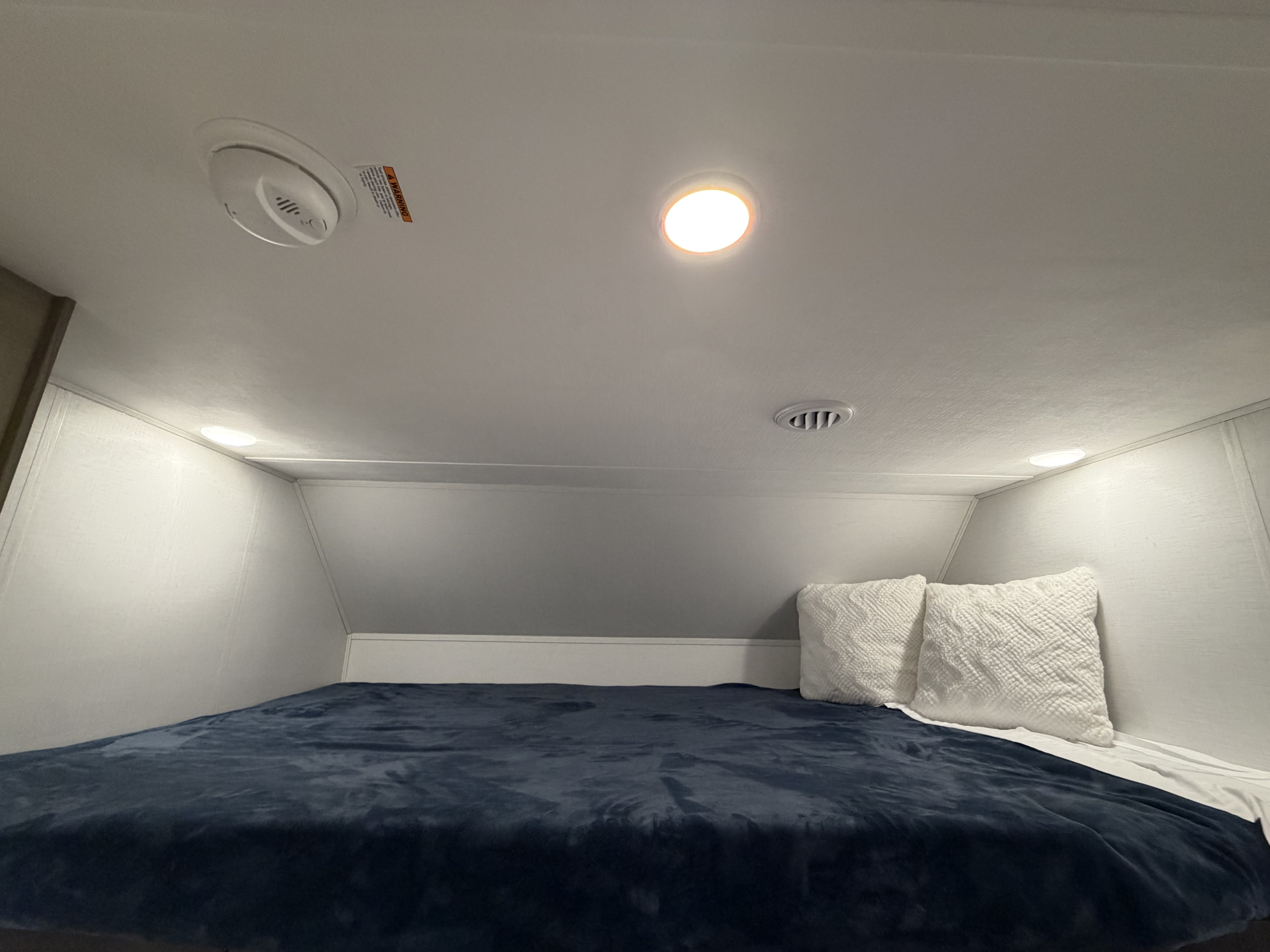 Cozy rv rental bunk: a dark blue bed with white pillows, overhead lighting, a smoke detector, and an air vent.. Forest River Sunseeker LE 2025