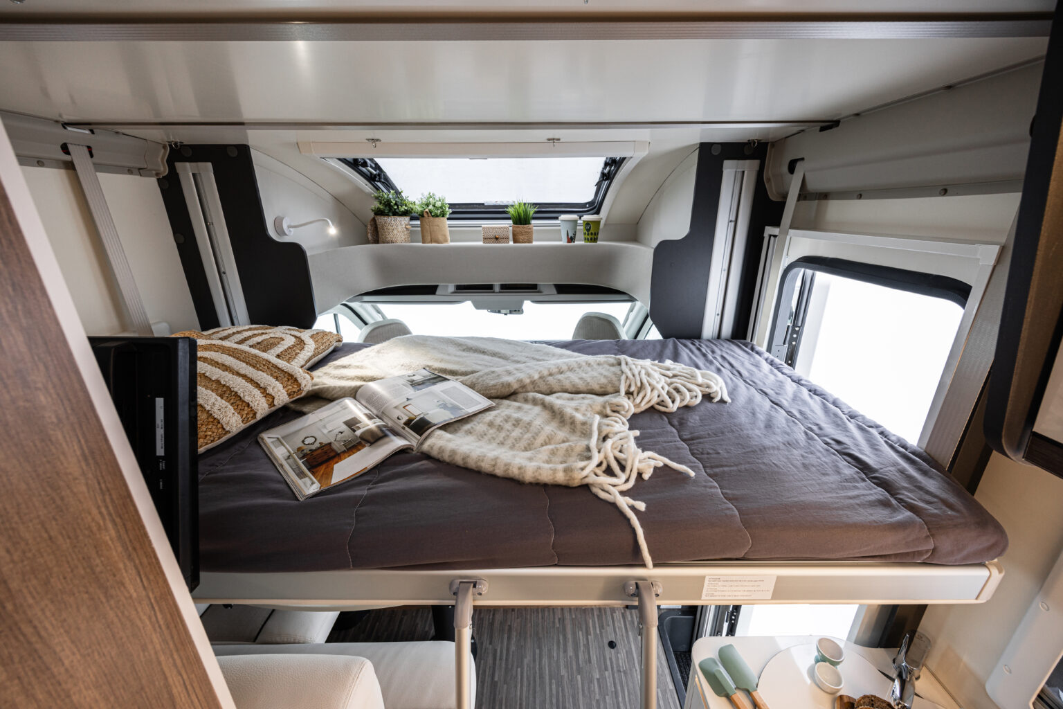 Comfortable drop-down bed in an RV rental, with a comforter, pillows, magazine, and plants creating a cozy sleeping space.. Elnagh baron 539 2023