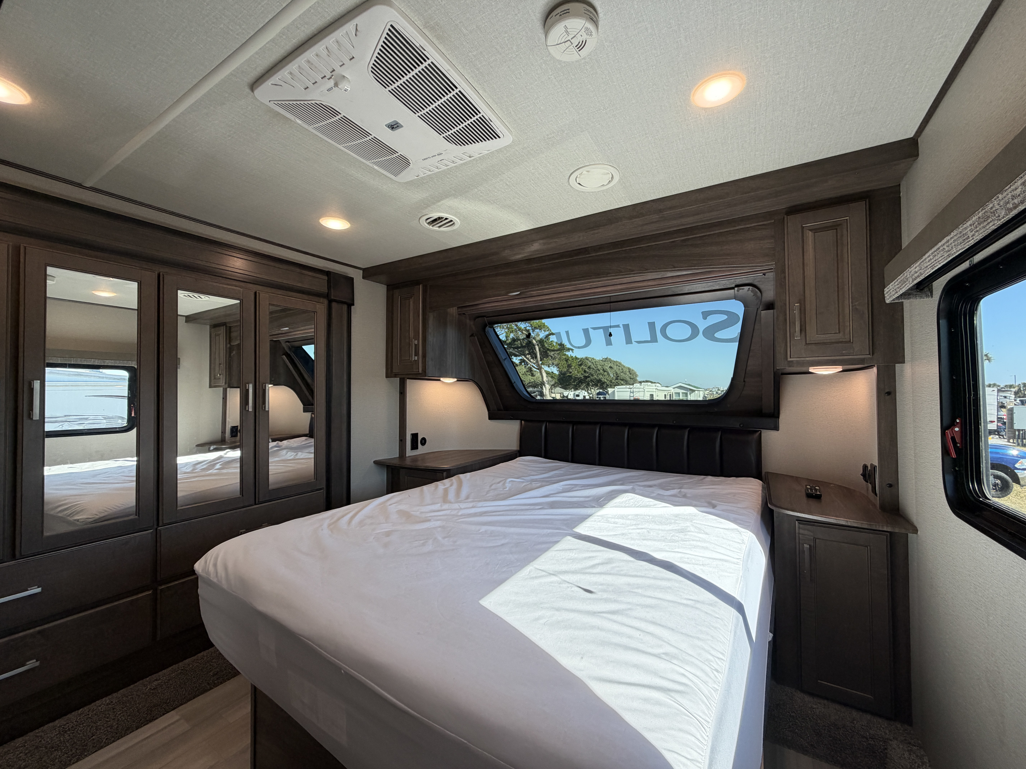 Modern RV rental bedroom featuring a comfy bed, dark wood storage, mirrored wardrobe, AC, and a scenic window view.. Grand Design Solitude 2021