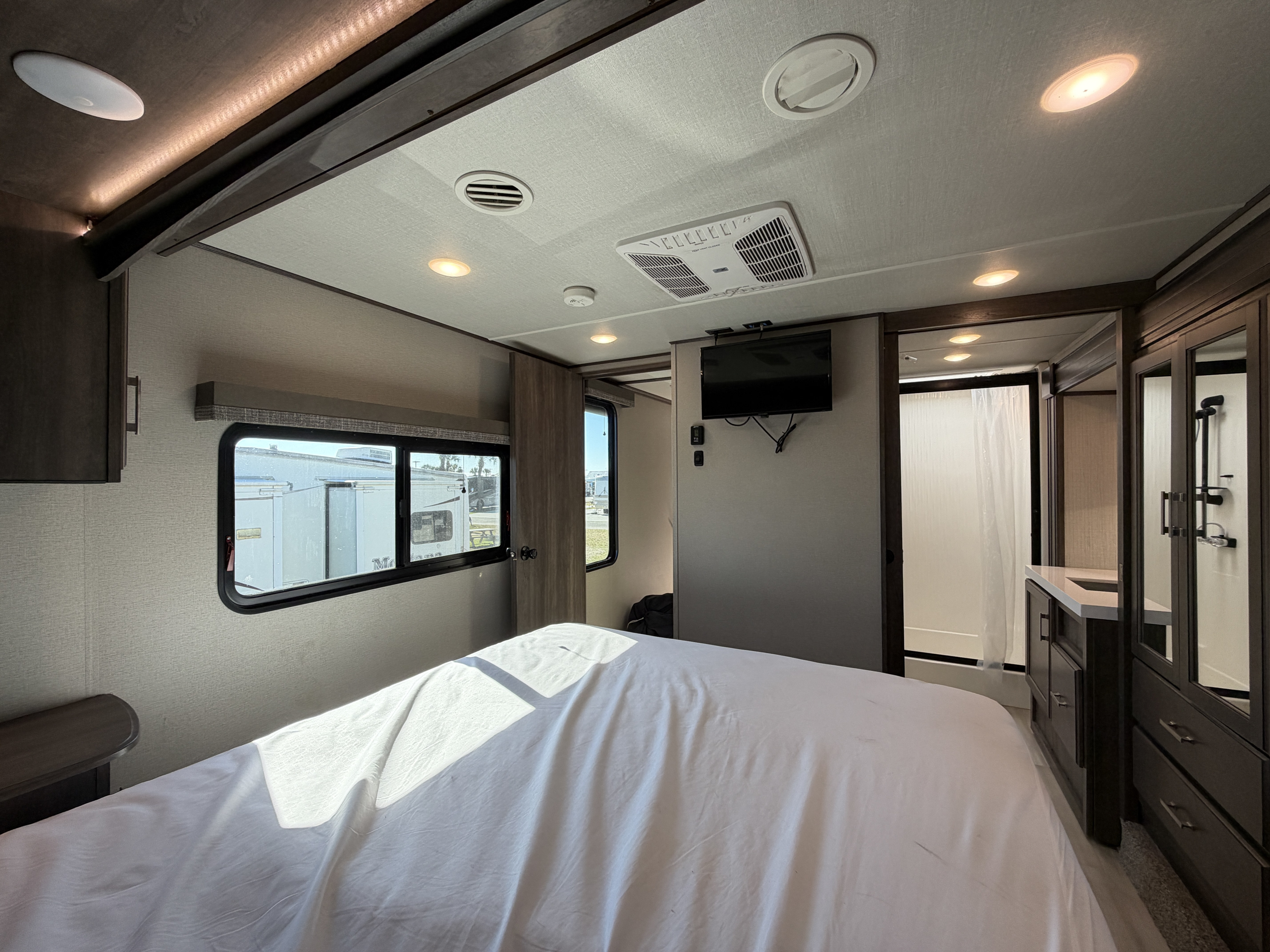 Interior of an RV rental bedroom with a large bed, sunlit windows, wall-mounted TV, and a glimpse of the ensuite bathroom.. Grand Design Solitude 2021
