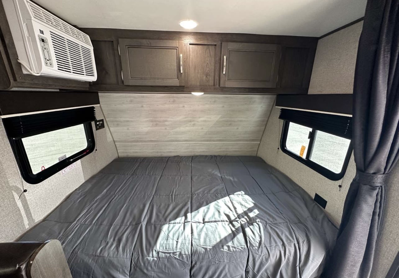 Cozy RV rental bedroom featuring a grey bed, overhead storage, side windows, and a wall-mounted AC unit.. Jayco Jay Flight Baja Edition 2023