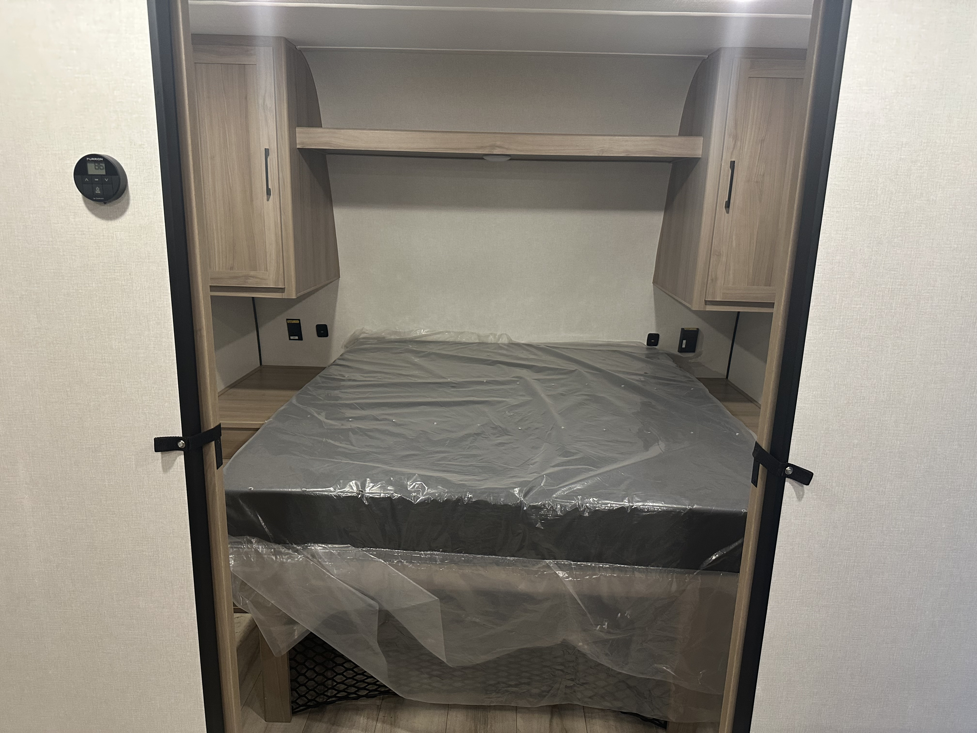 Inside an RV rental bedroom: a new, plastic-wrapped bed, overhead cabinets, and a thermostat. Ready for your journey.. Dutchmen Coleman Light 2025