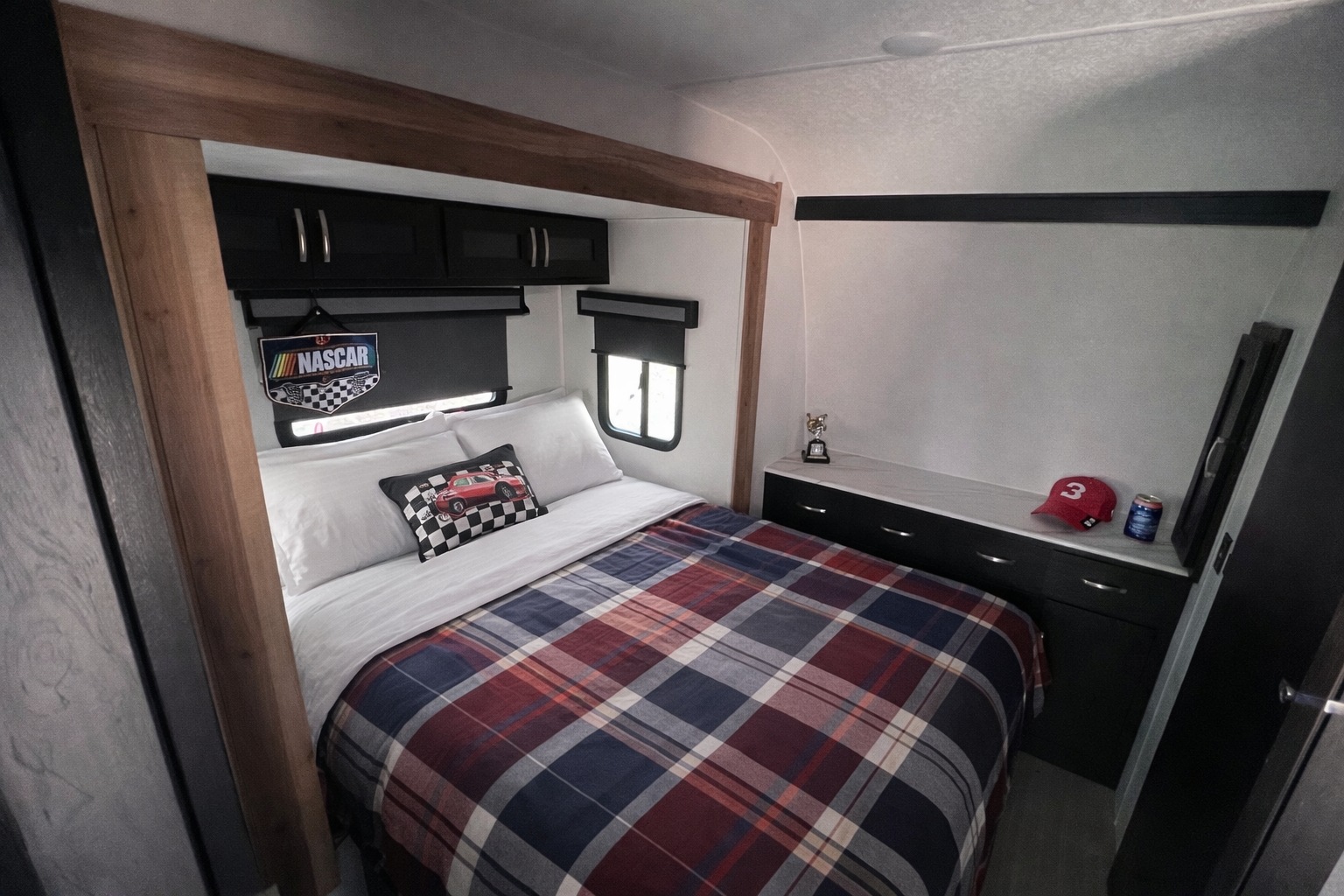 Cozy RV rental bedroom featuring a plaid bed, NASCAR decor, storage, and windows. Ready for your next adventure.. Work and Play Toy Hauler 2024