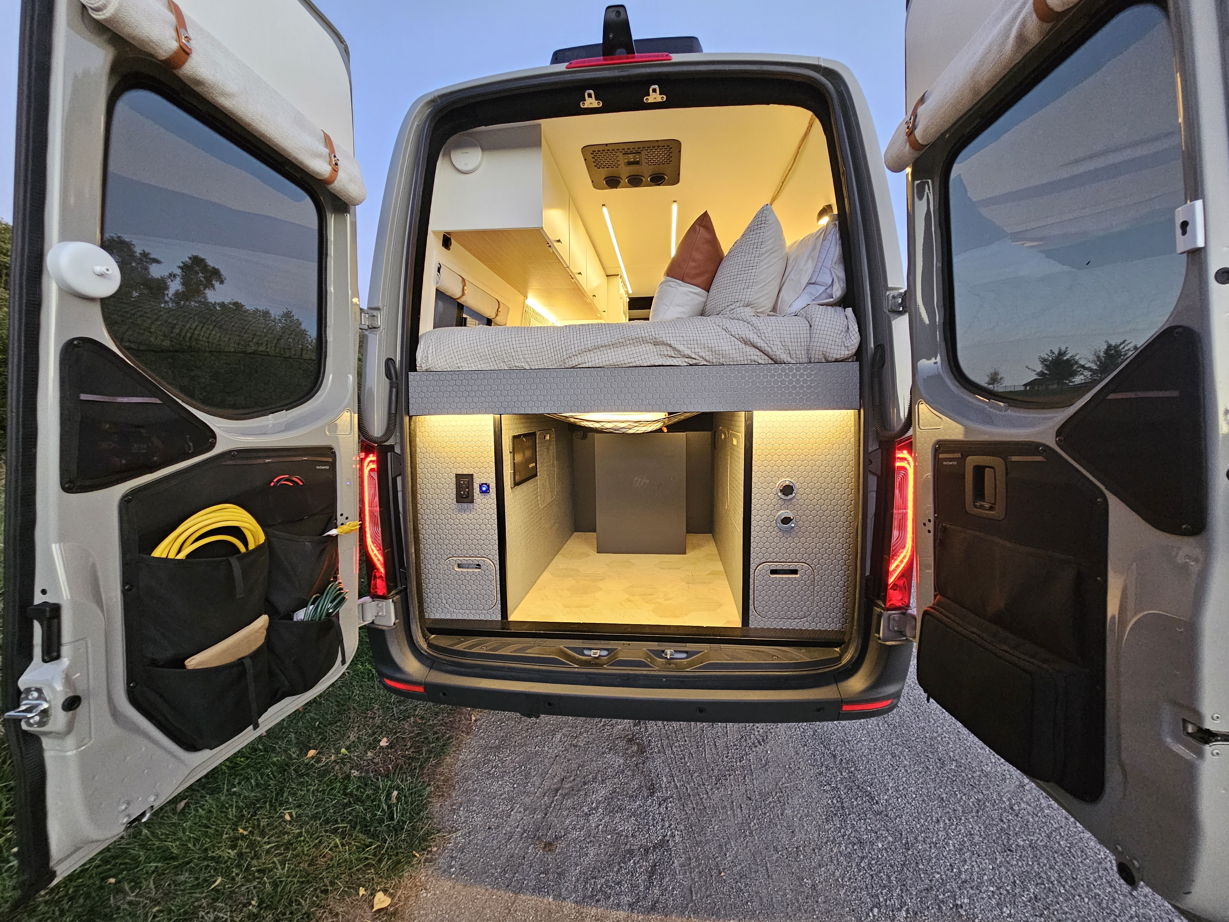 Experience an rv rental! Rear view of a custom camper van's open doors, revealing a comfy bed and illuminated storage area.. Mercedes-Benz Sprinter 2025