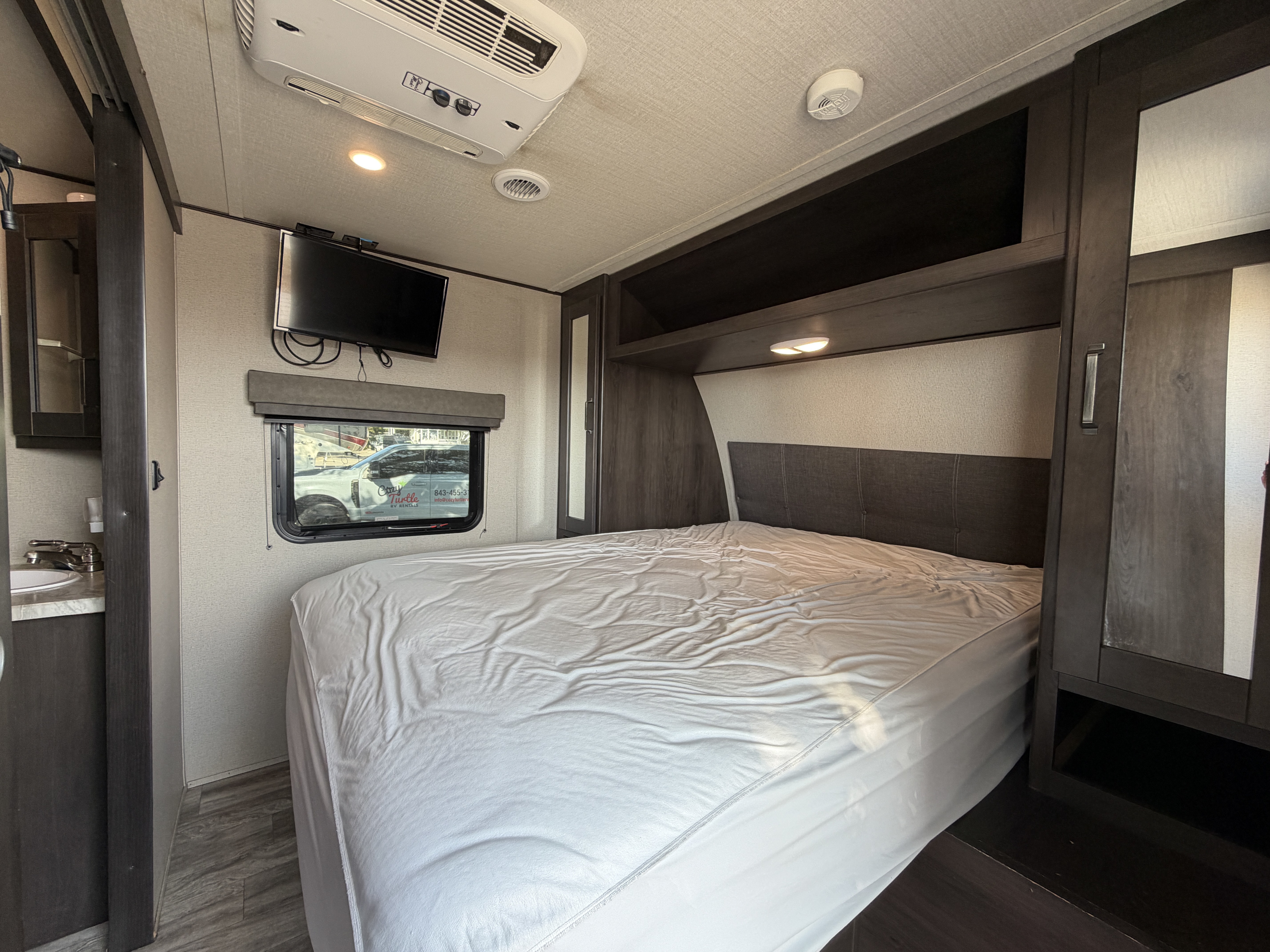 RV rental bedroom interior: features a comfortable bed, wall-mounted TV, window, AC unit, and a glimpse of a sink area.. Grand Design Transcend 2022