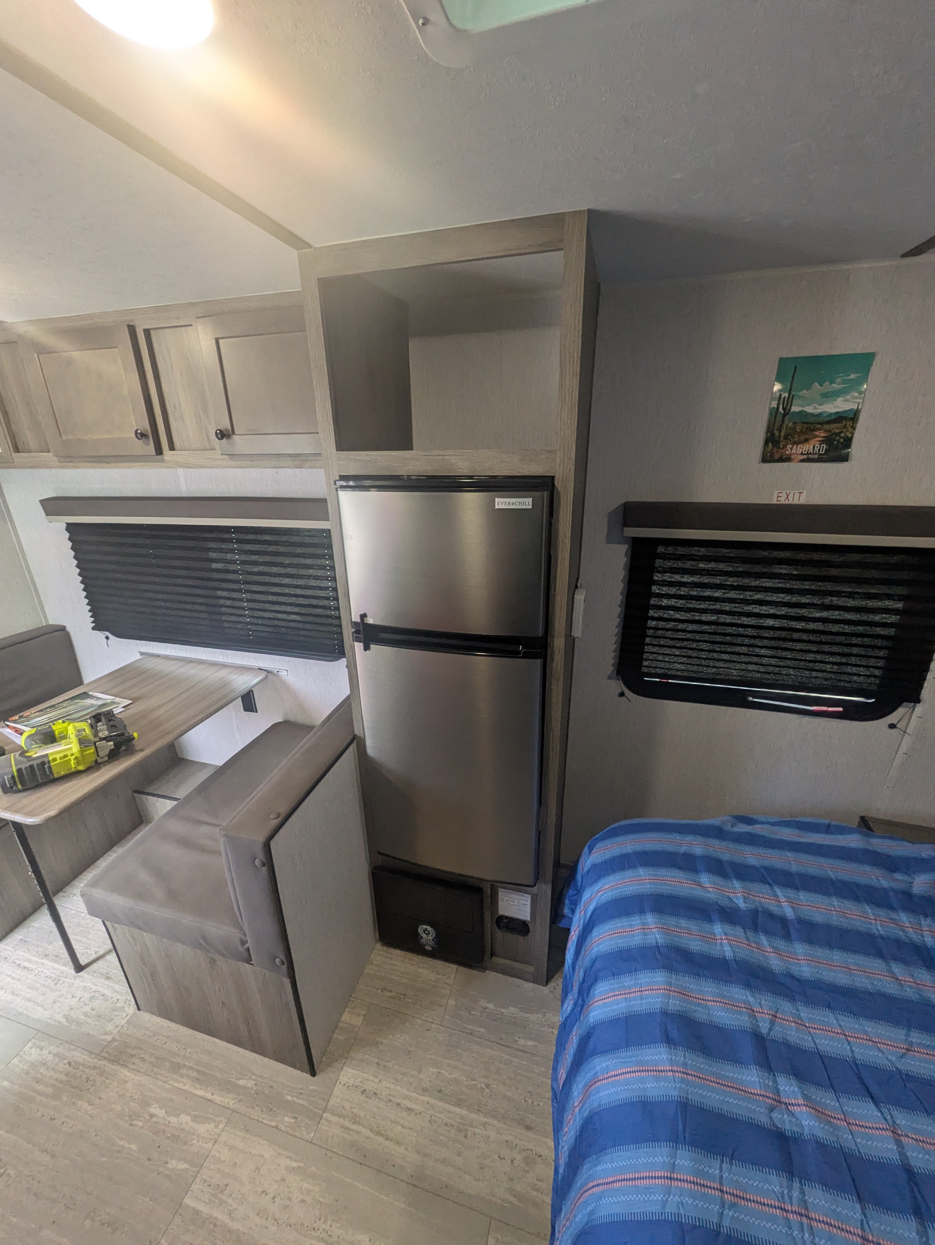 This RV rental features a compact kitchen/dinette area, fridge, storage, and a comfy bed with a striped comforter.. Sunset Park & Rv Inc. 19rb 2025