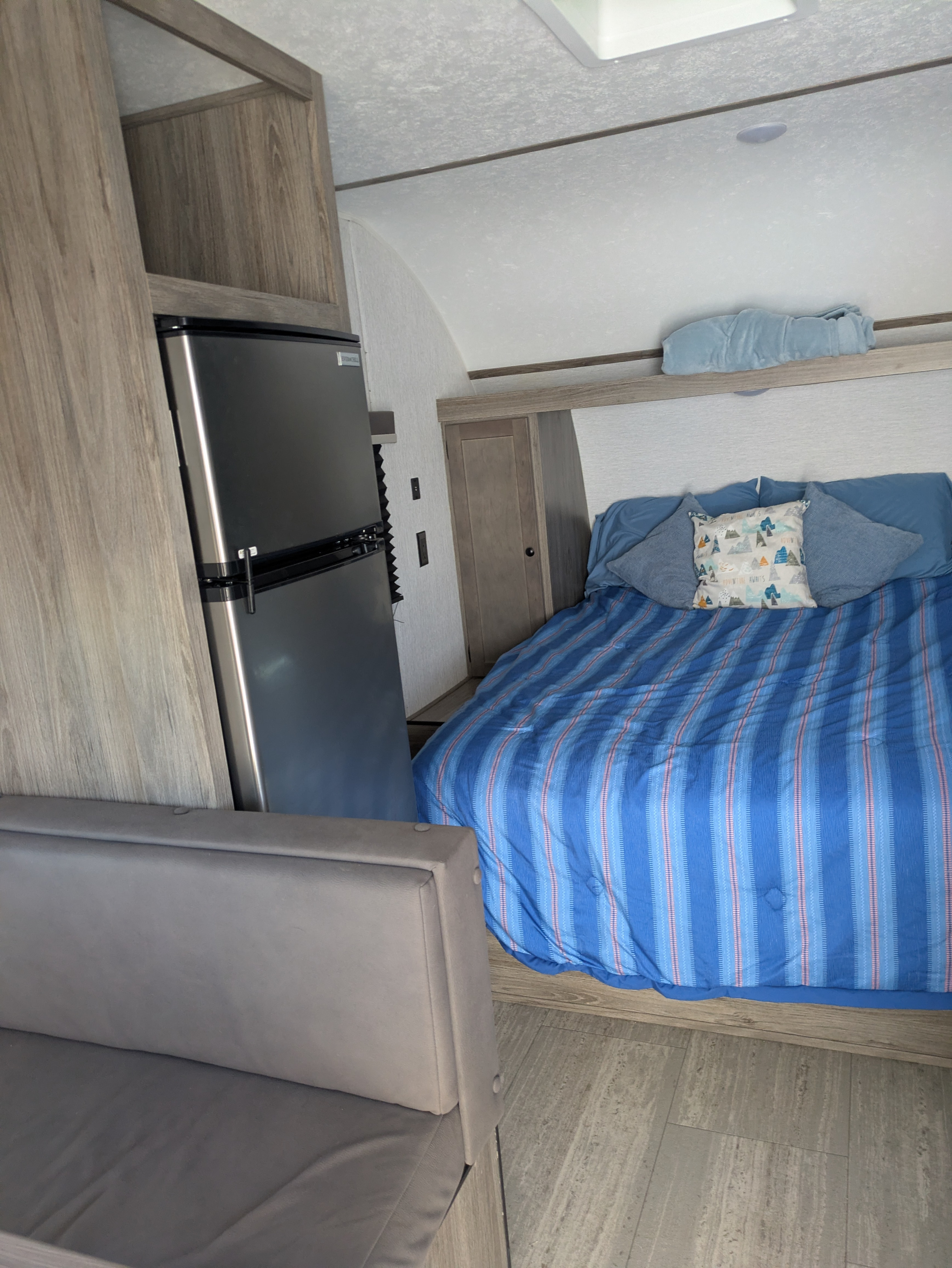 This cozy rv rental interior features a bed with blue bedding, a compact refrigerator, and a grey bench seat.. Sunset Park & Rv Inc. 19rb 2025