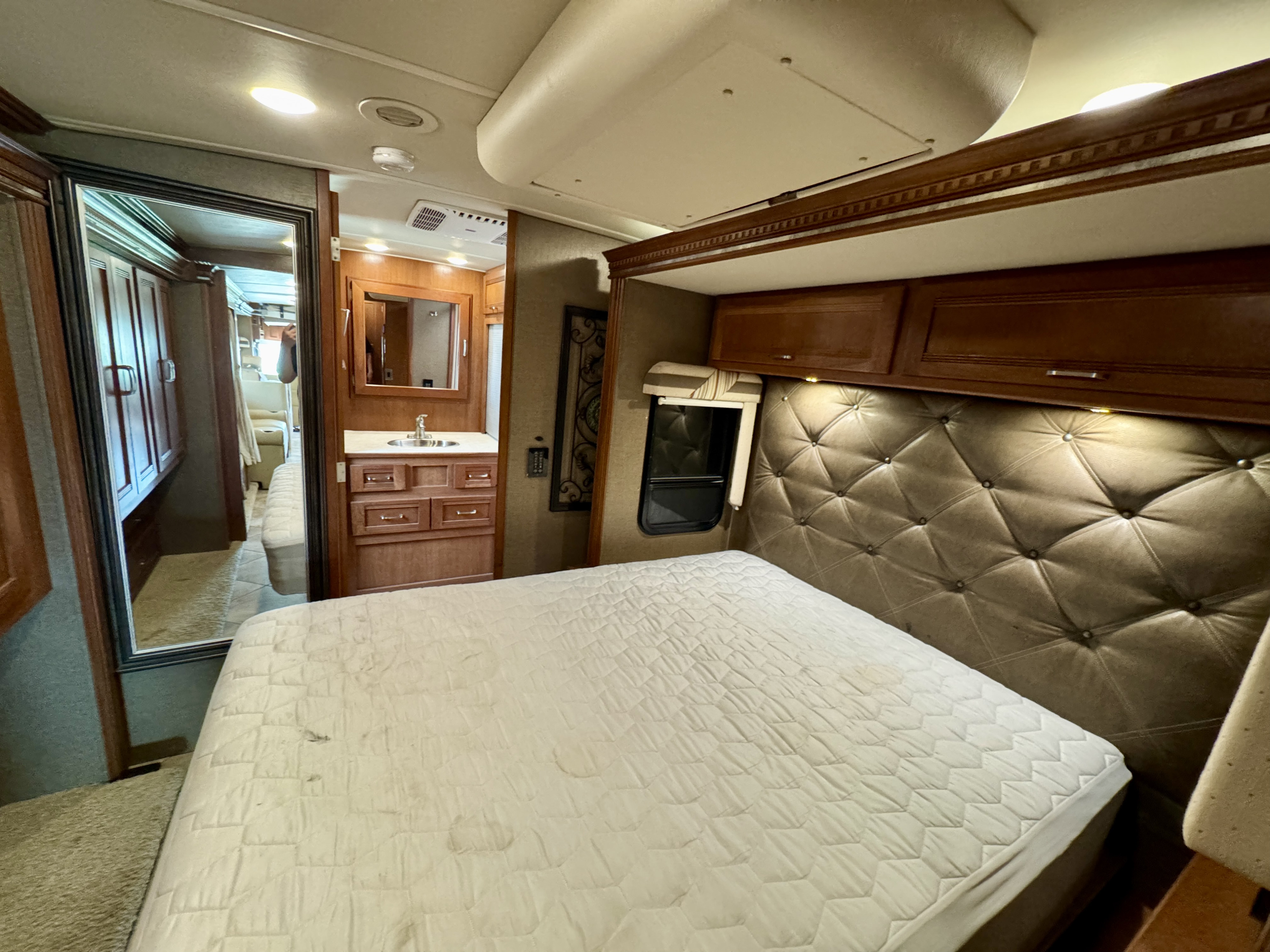 RV rental bedroom with a large bed, tufted headboard, overhead cabinets, and a mirror reflecting a vanity and hallway.. Winnebago Forza 2017