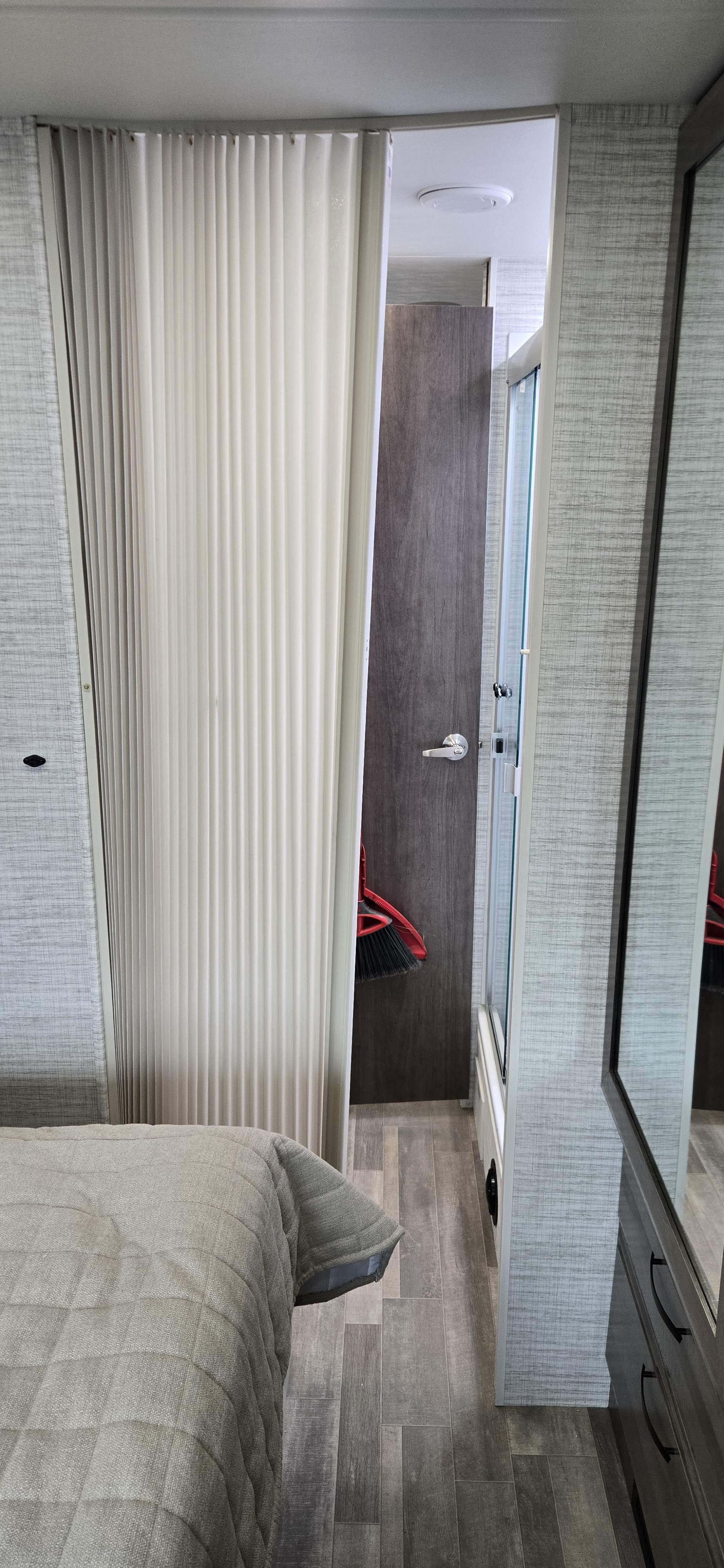 RV rental interior: bed, a pulled-back privacy curtain, dark wood door, and a shower with a glass door. A red dustpan is visible.. Jayco Greyhawk 2025