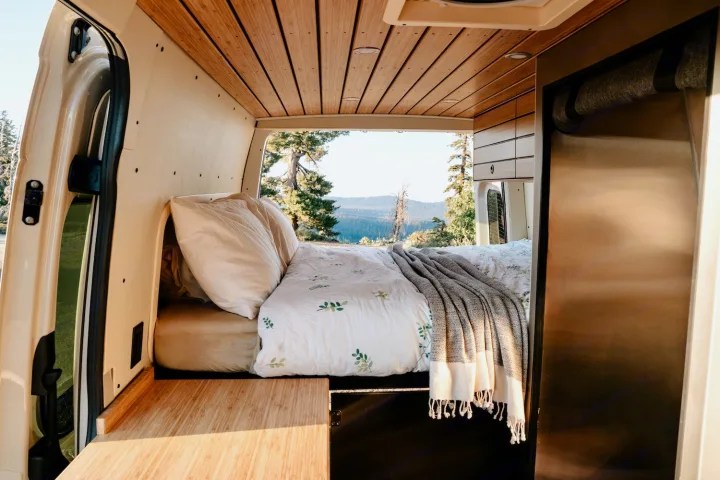 RV rental: Cozy camper van bedroom with a stunning mountain view from the back, featuring wood interior and a comfortable bed.. Mercedes Camper Van 2023