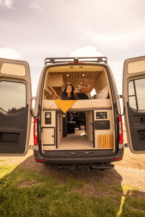 Relax in your spacious RV rental! A woman enjoys the custom camper van's cozy interior with rear doors open to nature.. Mercedes Benz Camper Van 2023