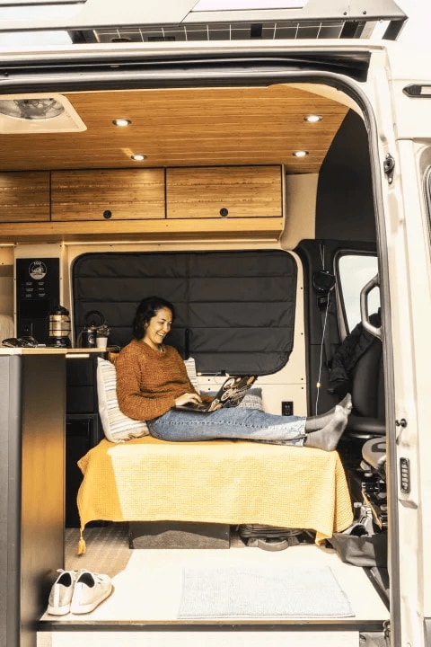 Experience an RV rental! A woman works on her laptop inside a bright, custom campervan with a cozy bed and wooden interior.. Mercedes Benz Camper Van 2023