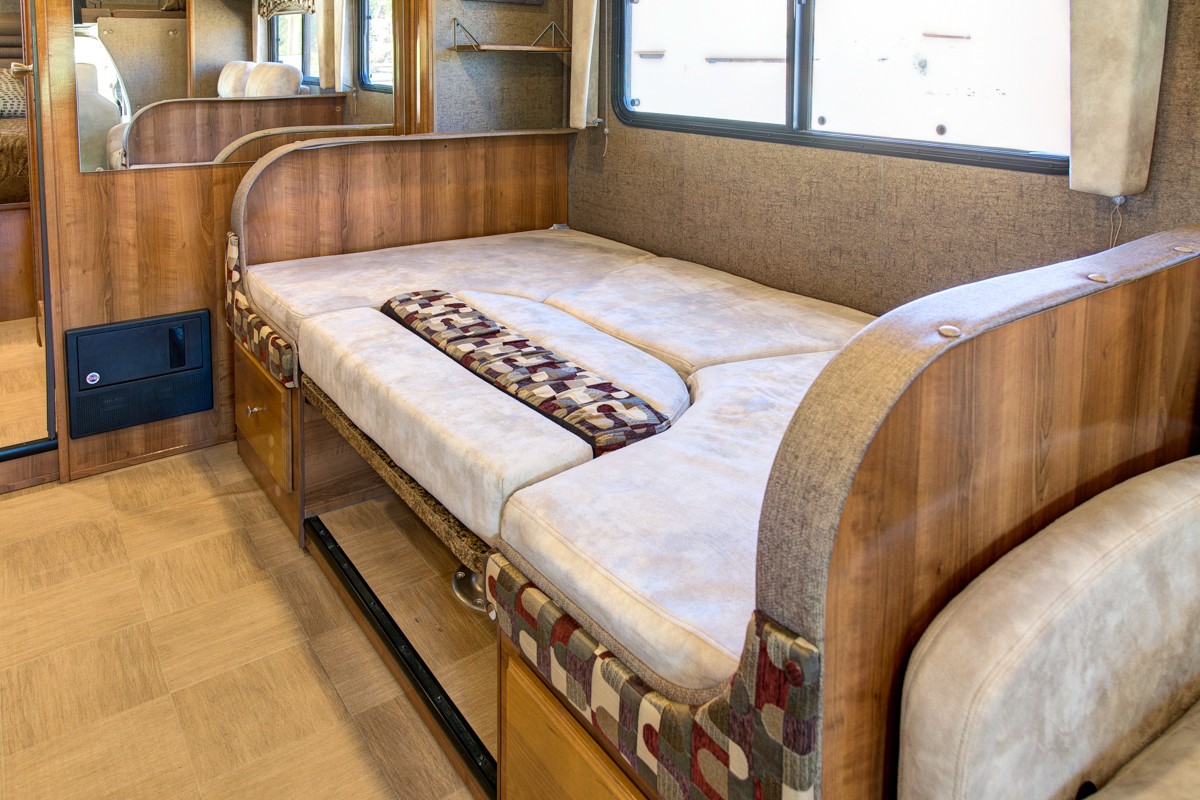RV rental interior featuring a cozy convertible dinette and bed setup with light wood, patterned cushions, and a window.. Forest River Coachmen Leprechaun 2016
