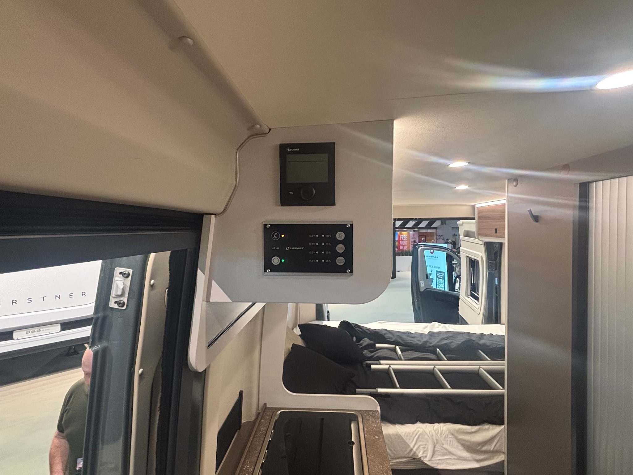 Inside a compact RV rental, showing the Truma and Lippert control panel, bed, and small kitchen area. Ideal for travel adventures.. Burstner B66 2026