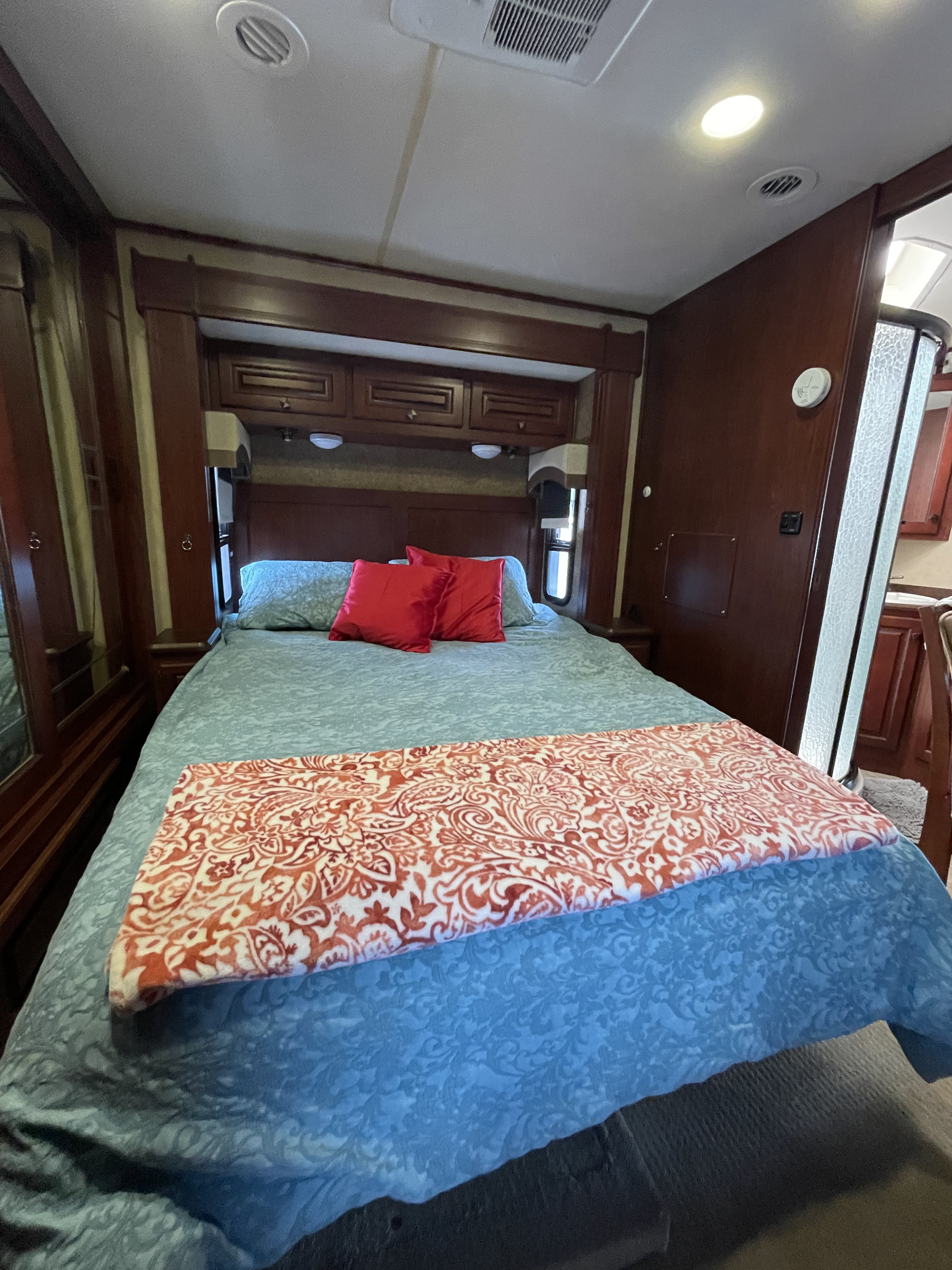 A cozy rv rental bedroom featuring a bed with blue patterned bedding, red pillows, a decorative blanket, and warm wooden cabinetry.. Berkshire Berkshire Motorhome 2013