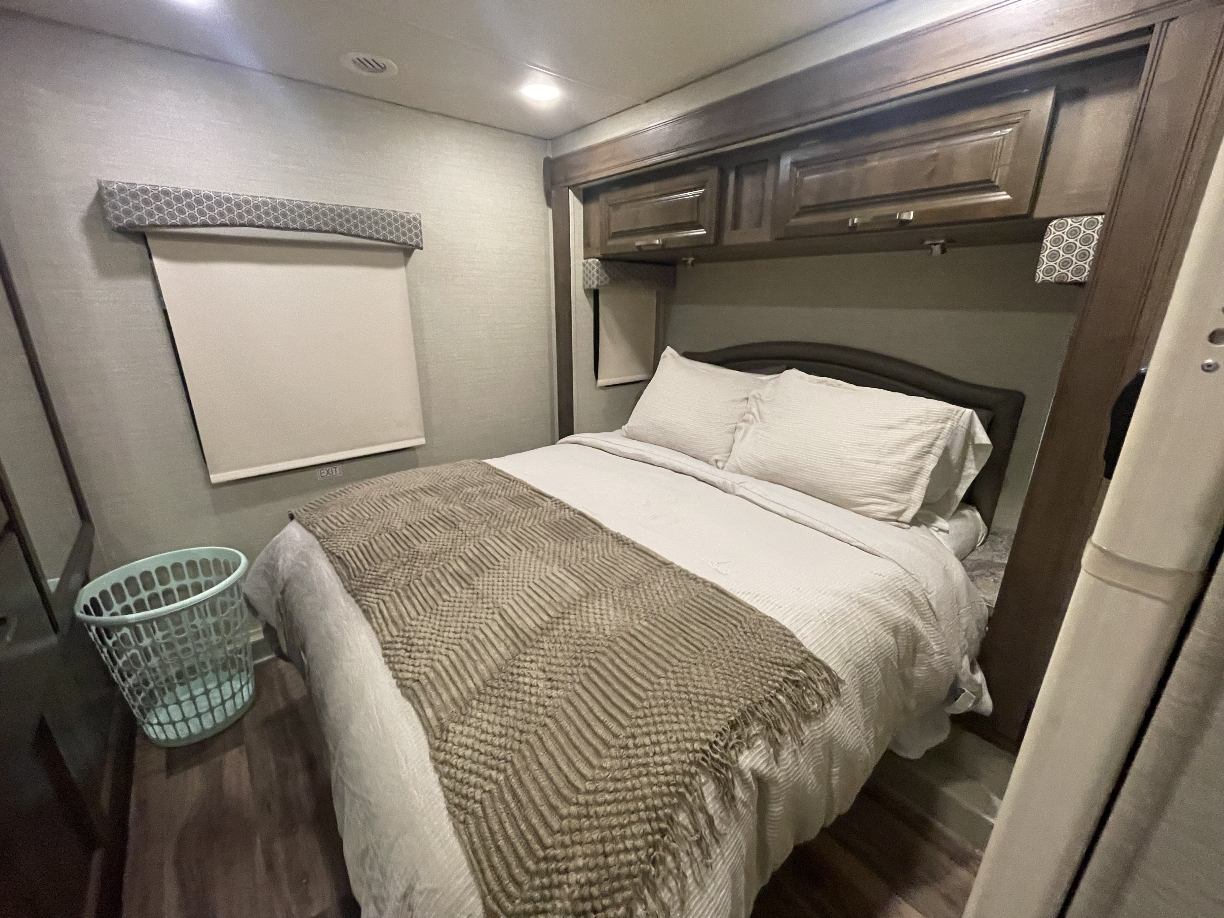 Inside an RV rental: a neatly made bed with white linens & a textured throw, overhead storage, window shade, & laundry basket.. Jayco Greyhawk 2019