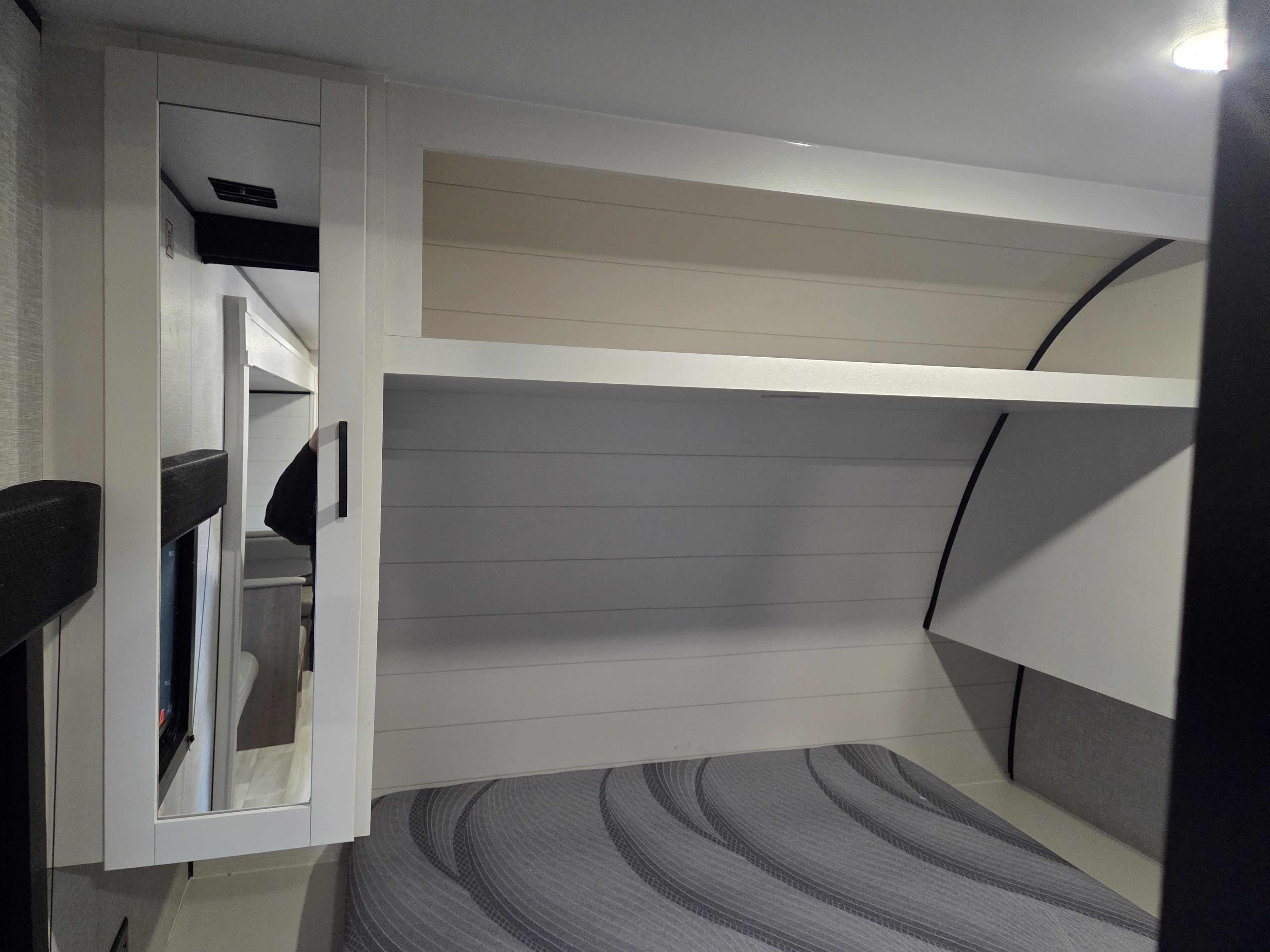 Compact RV rental bedroom featuring a grey bed, white shiplap walls, overhead shelf, and mirrored cabinet for storage.. Open Range Conventional 26BHS Travel Trailer 2025