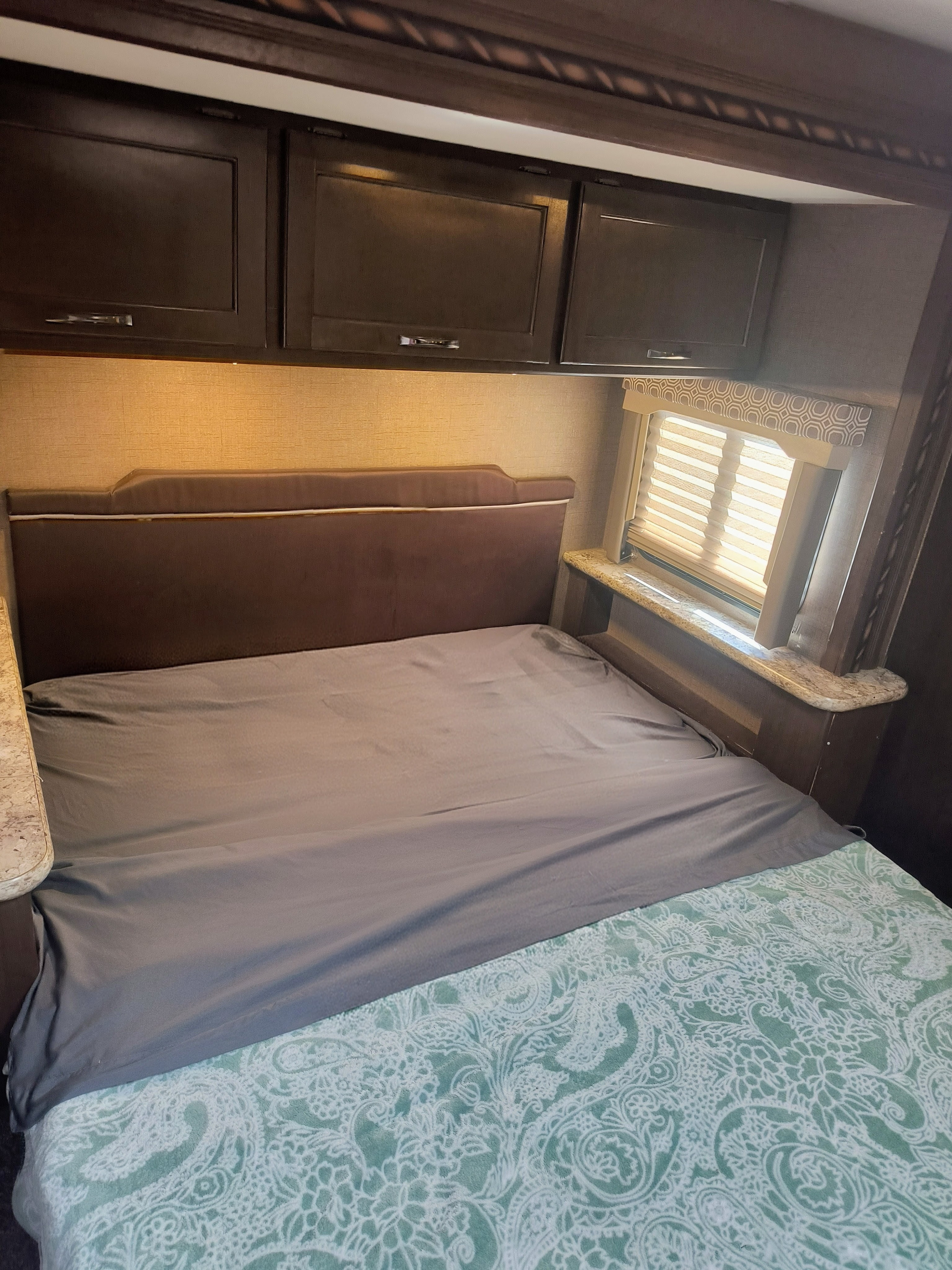 RV rental bedroom with a cozy bed, green patterned blanket, padded headboard, dark wood cabinets, and a window.. Thor Motor Coach Four Winds 2017