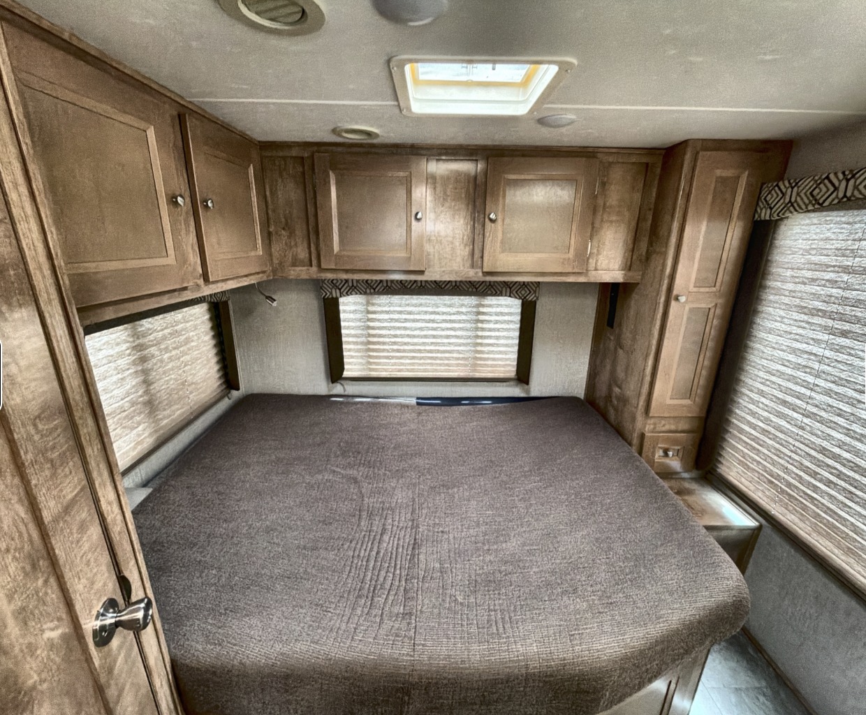 An RV rental's compact bedroom features a large bed, wooden storage cabinets, and window blinds.. Coachman Leprachaun 2020