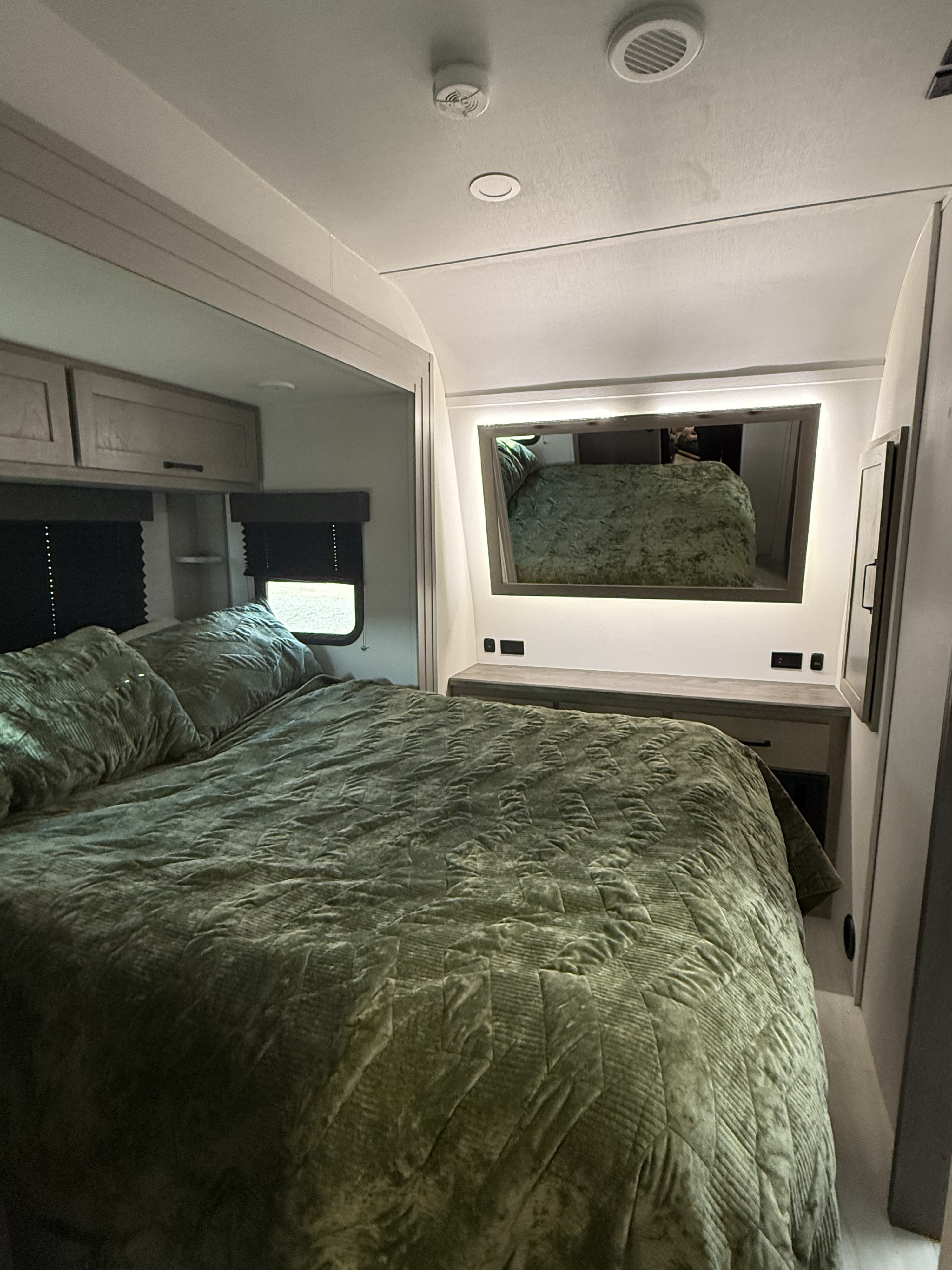 A modern RV rental bedroom featuring a comfortable green bed, overhead cabinets, and a stylish illuminated mirror.. Grand Design Momentum 2025