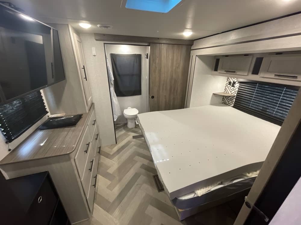 RV rental bedroom features a large bed, dresser, TV, and a small toilet area visible through the sliding door.. Crossroads RV Zinger 2024