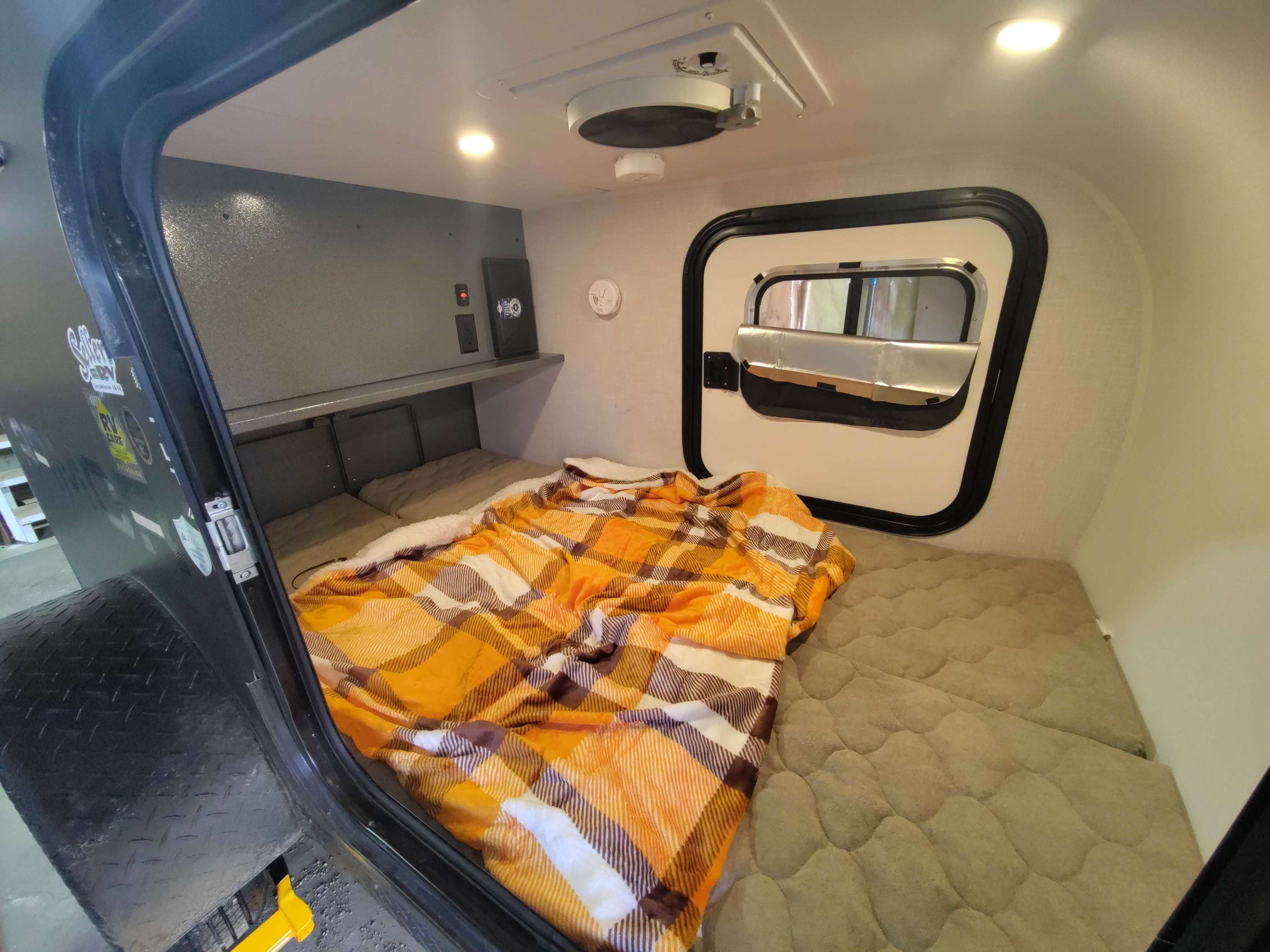 Inside this compact RV rental: A comfy bed with an orange plaid blanket, window, fan, and shelf for a restful journey.. Skinny Guy Campers Armor 2026
