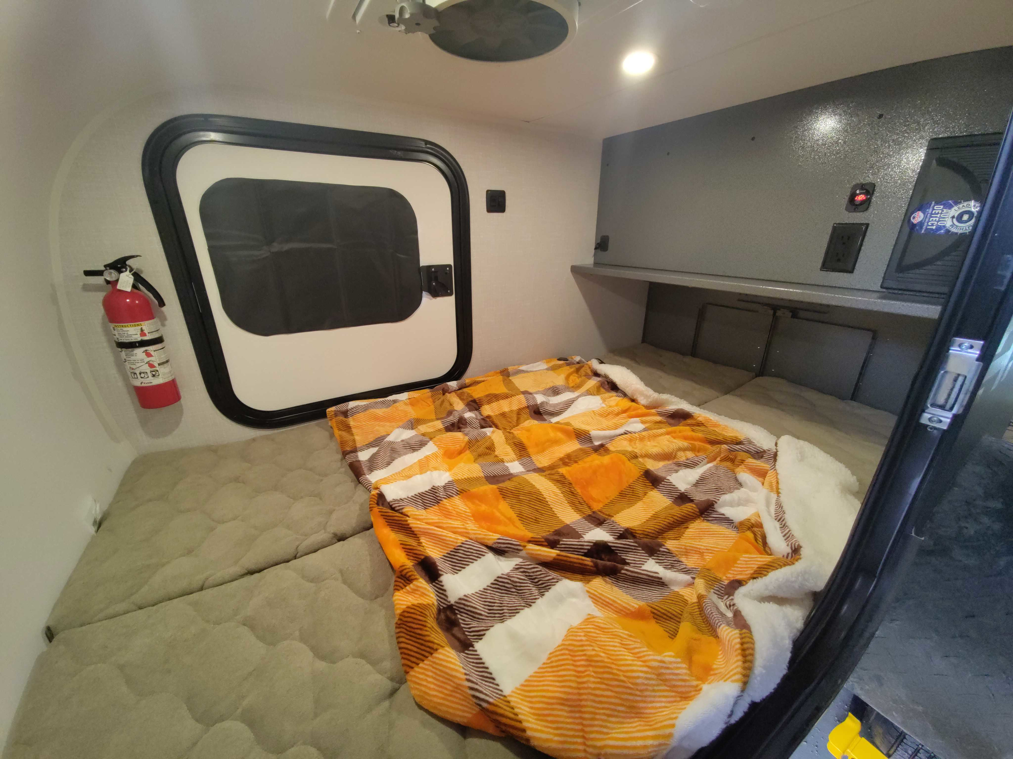 Inside a cozy **rv rental**: bed with plaid blanket, fire extinguisher, overhead fan, and shelf with power. Ideal for compact trips.. Skinny Guy Campers Armor 2026