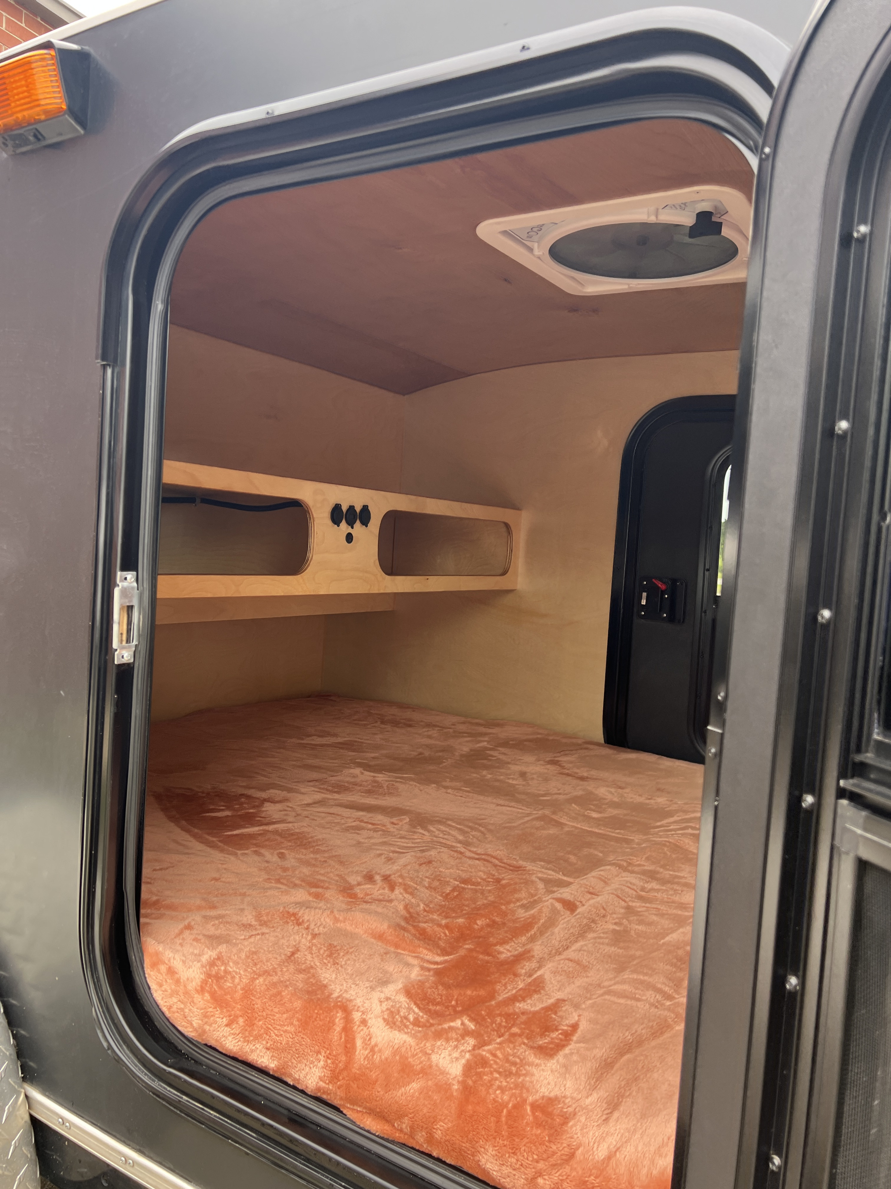 Welcoming RV rental interior: orange bed, wood-paneled walls/ceiling, shelf with outlets, and open door revealing the exterior.. Wander Tears Renegade LT 2021