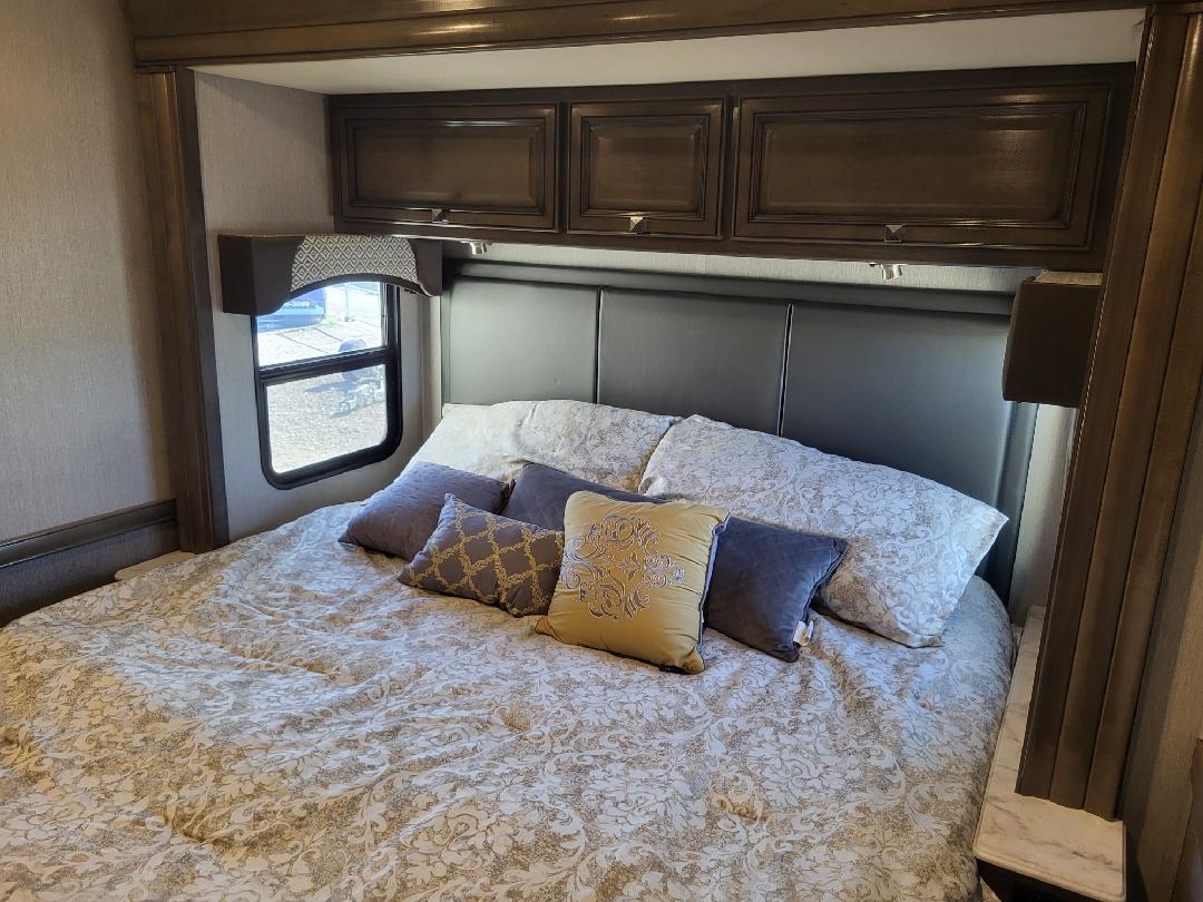 Cozy RV rental bedroom featuring a large bed with elegant patterned bedding, various pillows, overhead cabinets, and a side window.. Fleetwood Fortis 2020