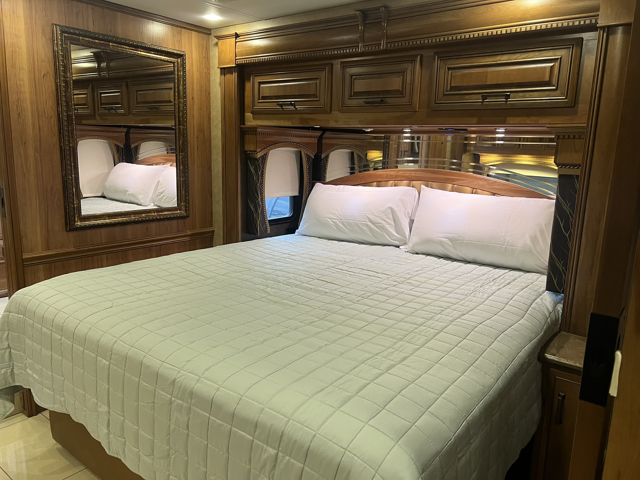 Cozy RV rental bedroom: large bed with green quilted spread, rich wooden cabinetry, mirrored headboard, and decorative wall mirror.. Entegra Coach Aspire 2015