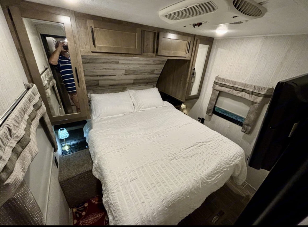 RV rental bedroom: a cozy space with white bed, wood cabinets, AC, and a person reflected in the mirror. Compact travel comfort.. Forest River Palomino Puma 2021