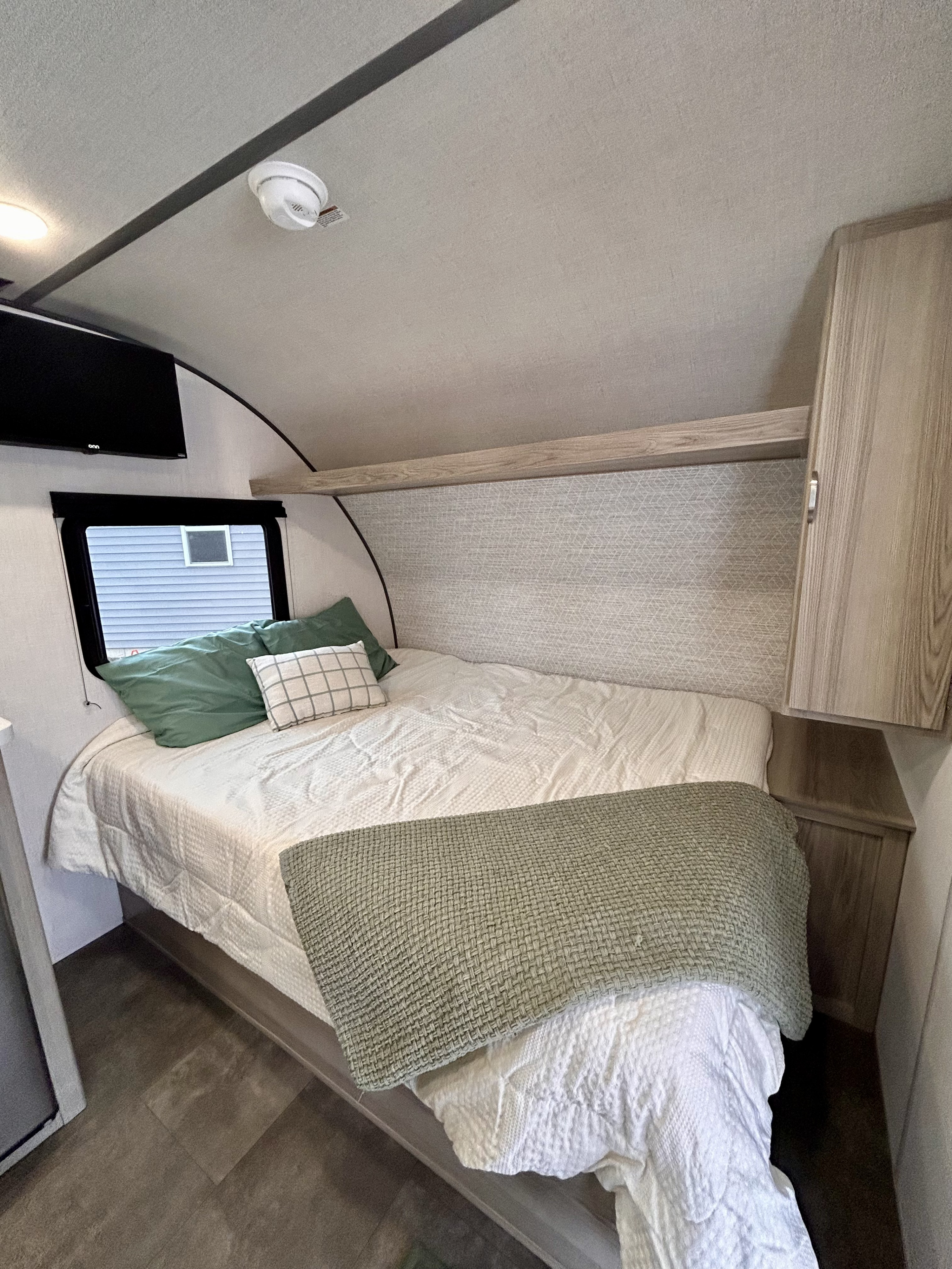 RV rental interior: bedroom with a bed, green pillows, white comforter, wall-mounted TV, and ceiling smoke detector visible.. Forest River Wildwood FSX 2025