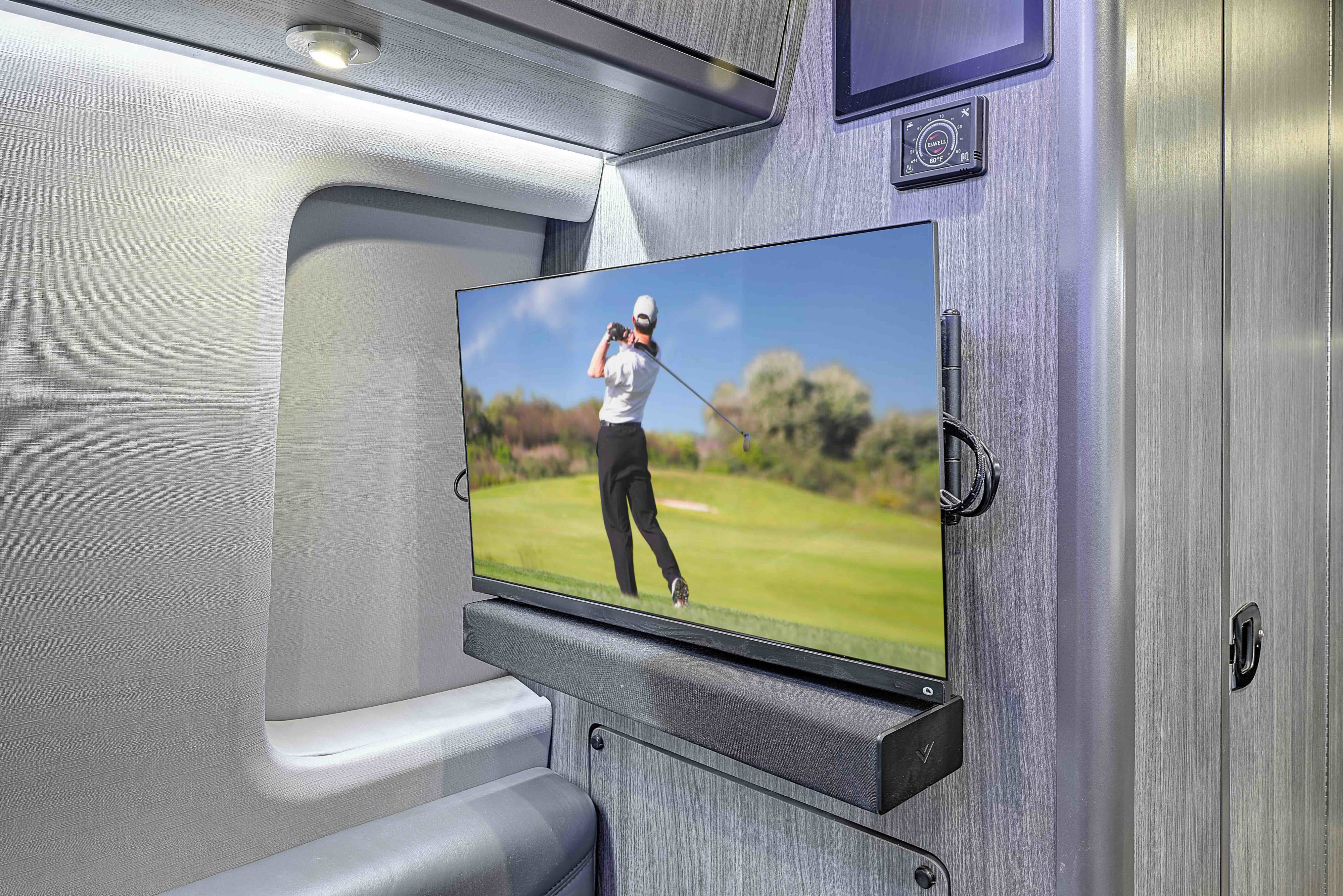 Modern RV rental interior with a wall-mounted TV showing a golfer, soundbar, and climate controls for comfort.. Grech Strada Ion Mercedes Sprinter 2025