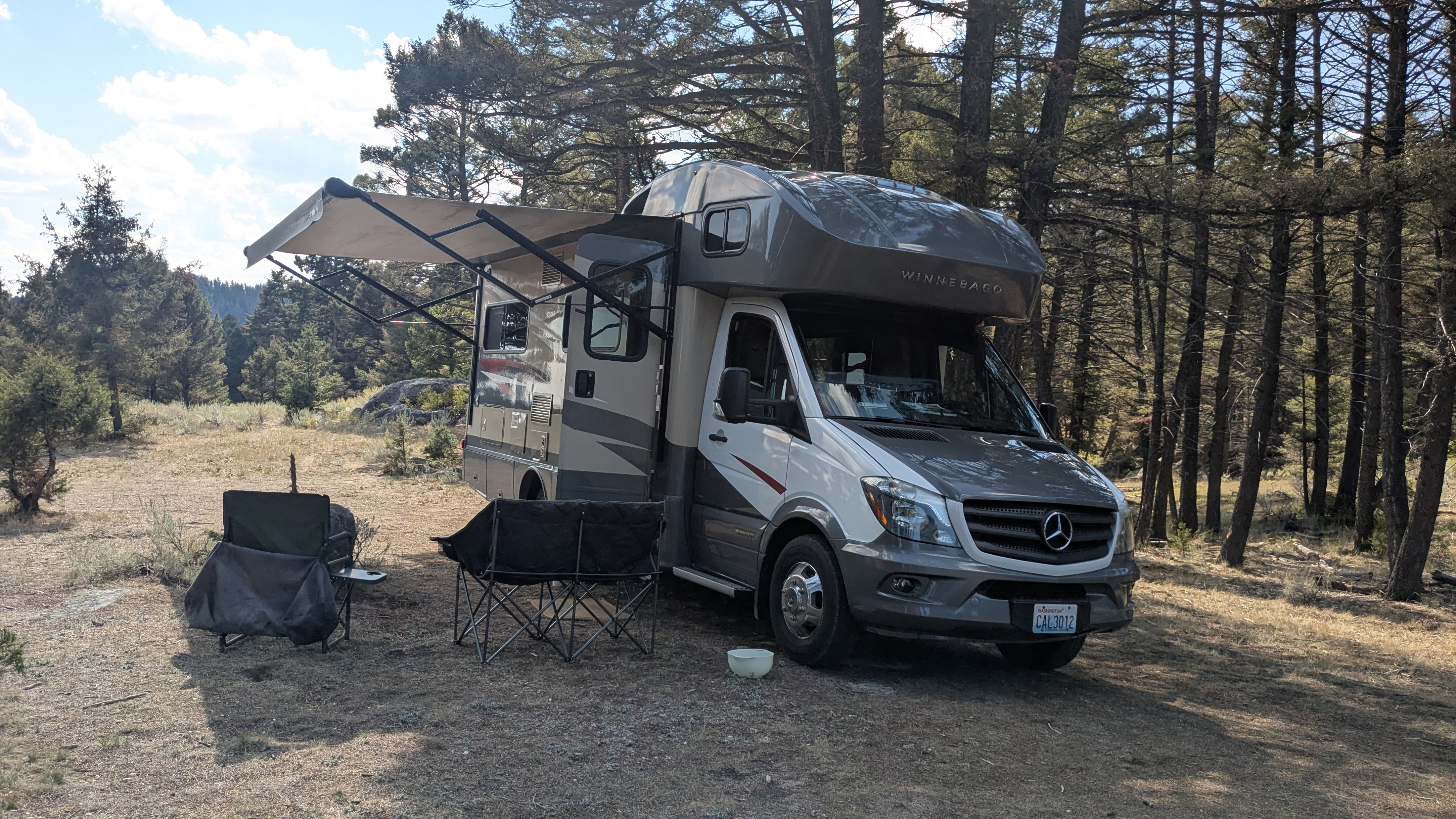 This Winnebago rv rental with an extended awning and chairs sits in a sunny, forested campsite, ready for outdoor adventure.. Winnebago View 2017