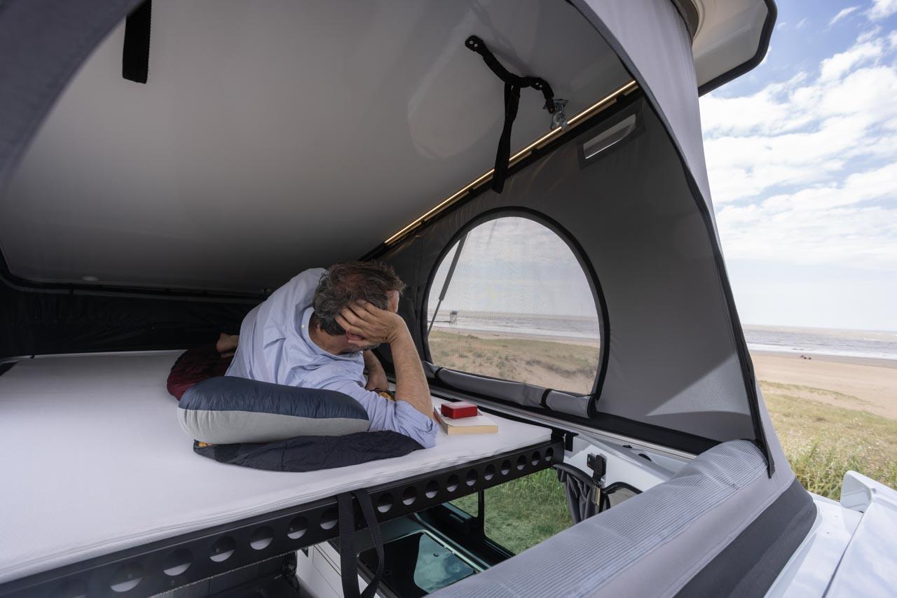 A man relaxes inside a comfy rv rental pop-top camper, enjoying beach and ocean views from his bed.. Handroad 2026 2026