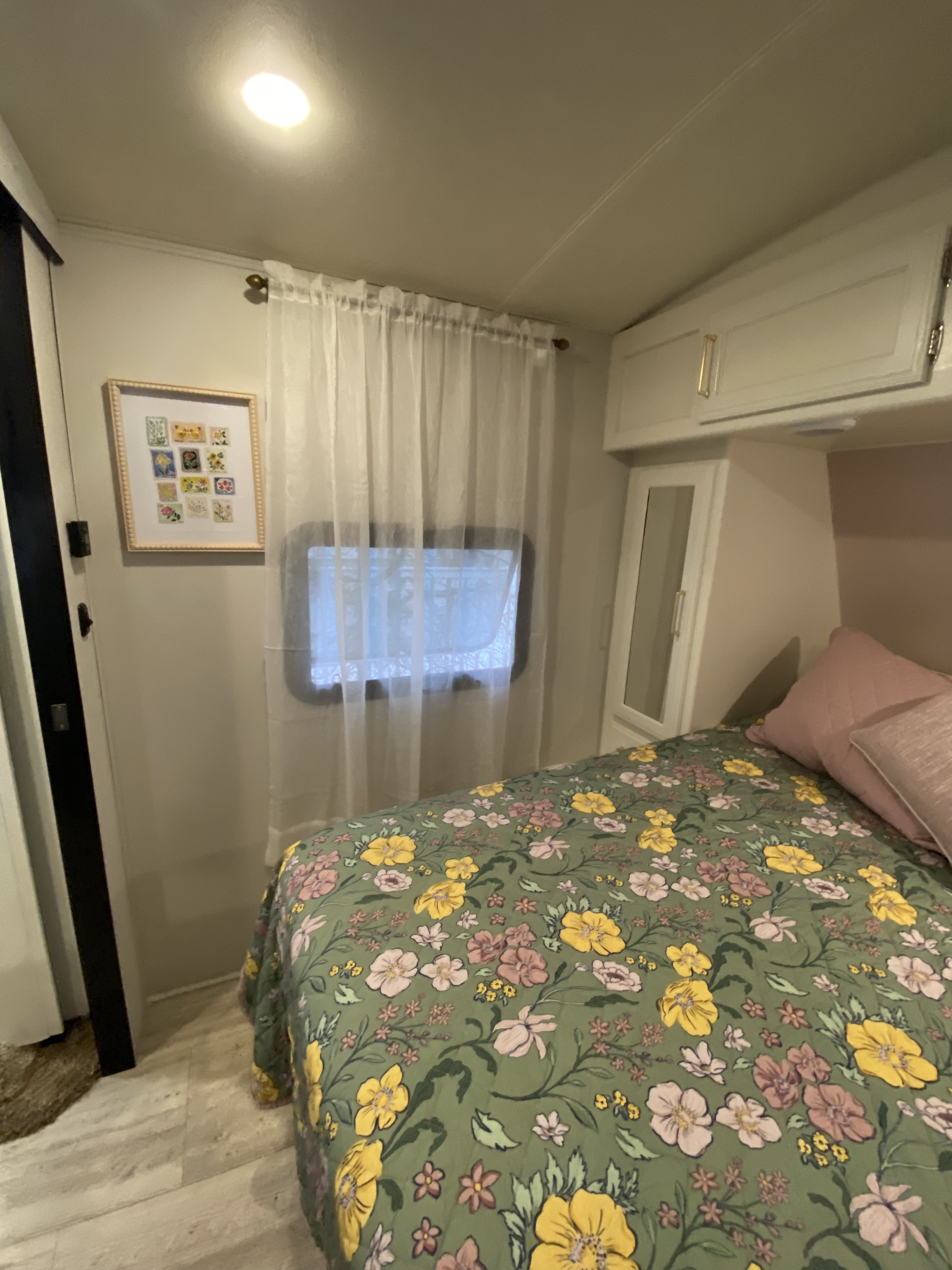Inside an RV rental, a cozy bedroom with a green floral comforter, sheer window curtains, and convenient overhead storage.. Grand Design Imagine 2017