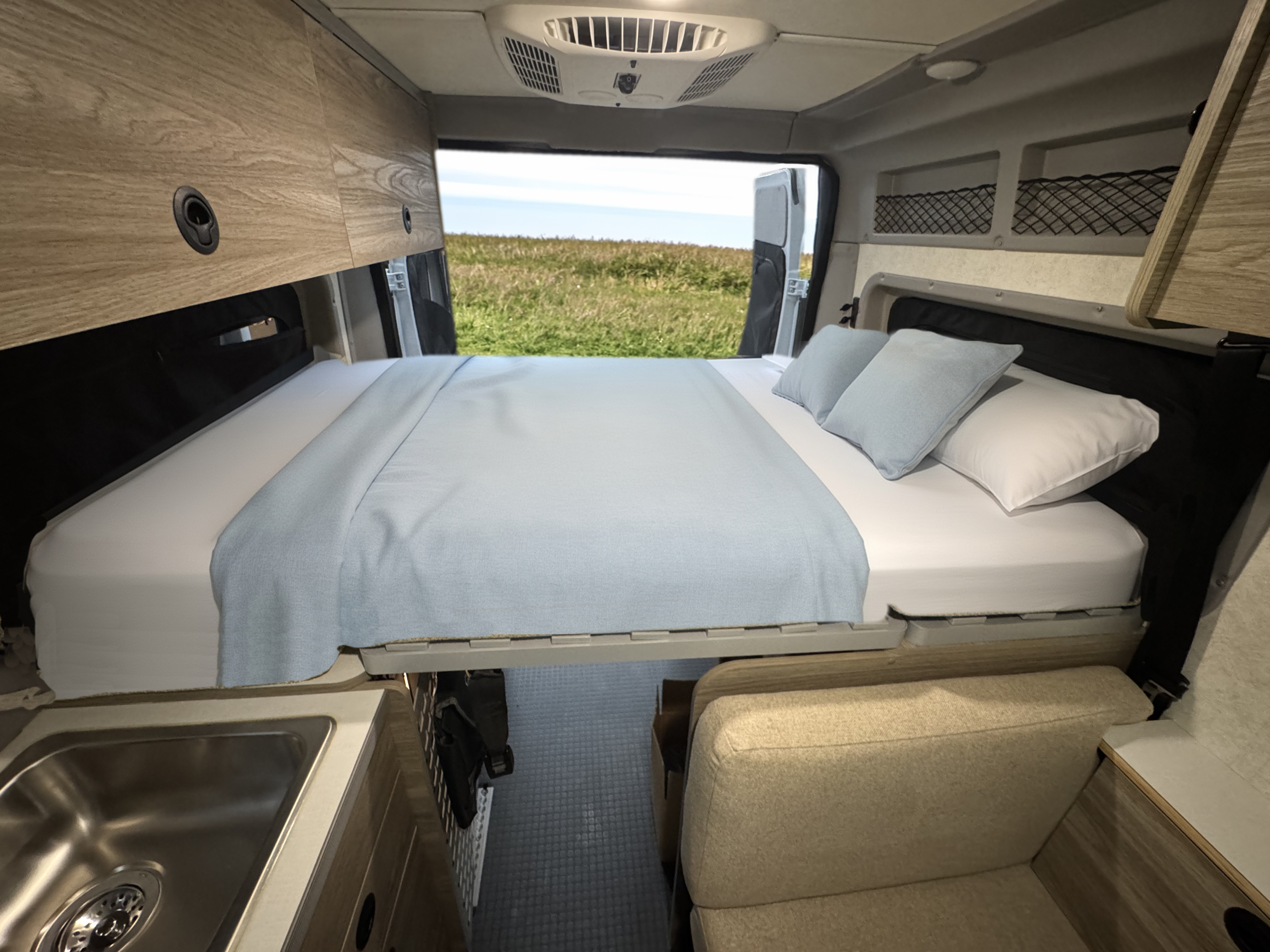 Inside a bright rv rental, a made bed with a blue blanket, overhead storage, a sink, and a clear view to green outdoors.. Winnebago Solis 2025