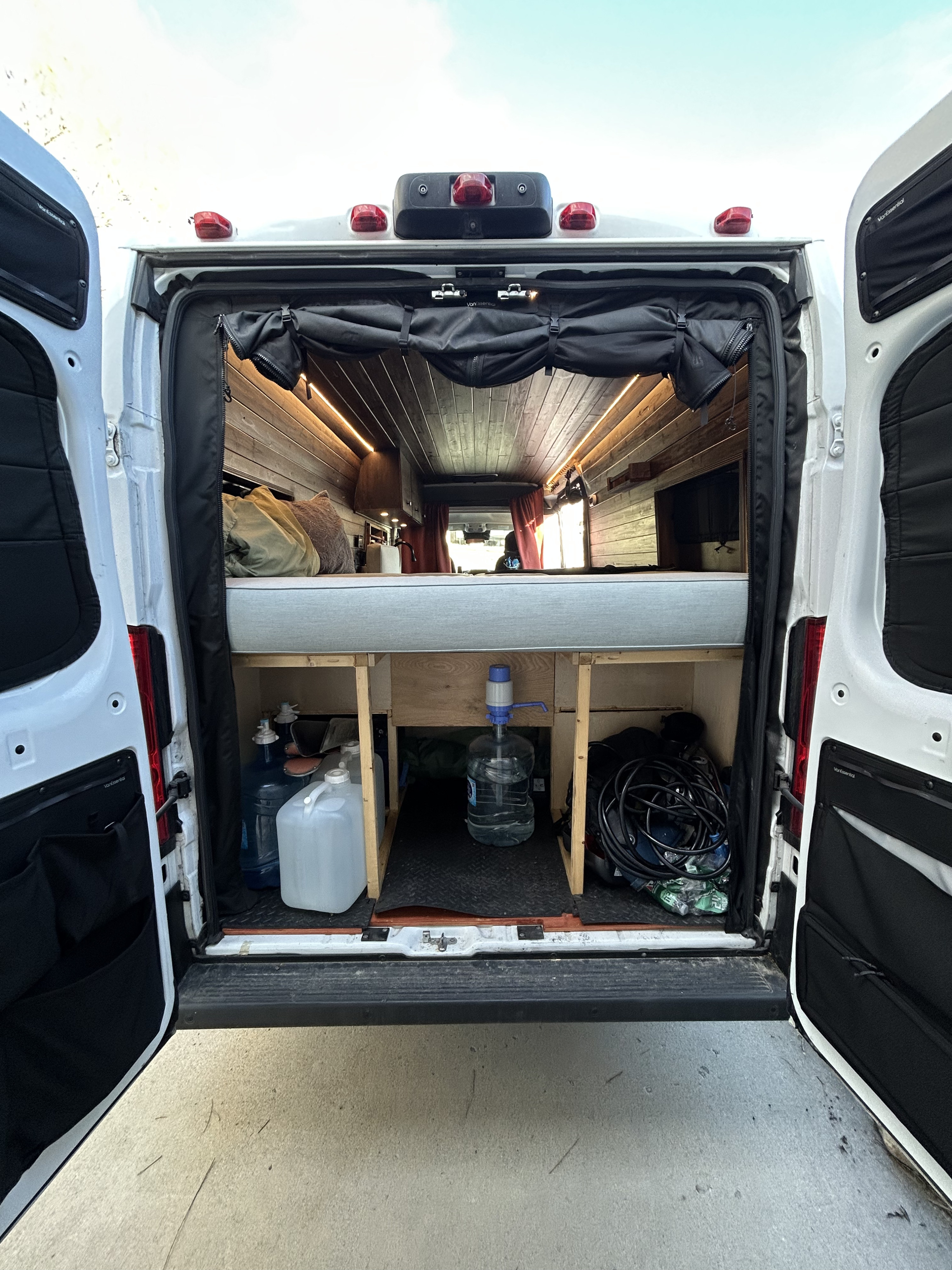 RV rental: Inside a camper van, showing a cozy bed, under-bed storage with water jugs, and wooden paneling.. RAM Promaster 2022